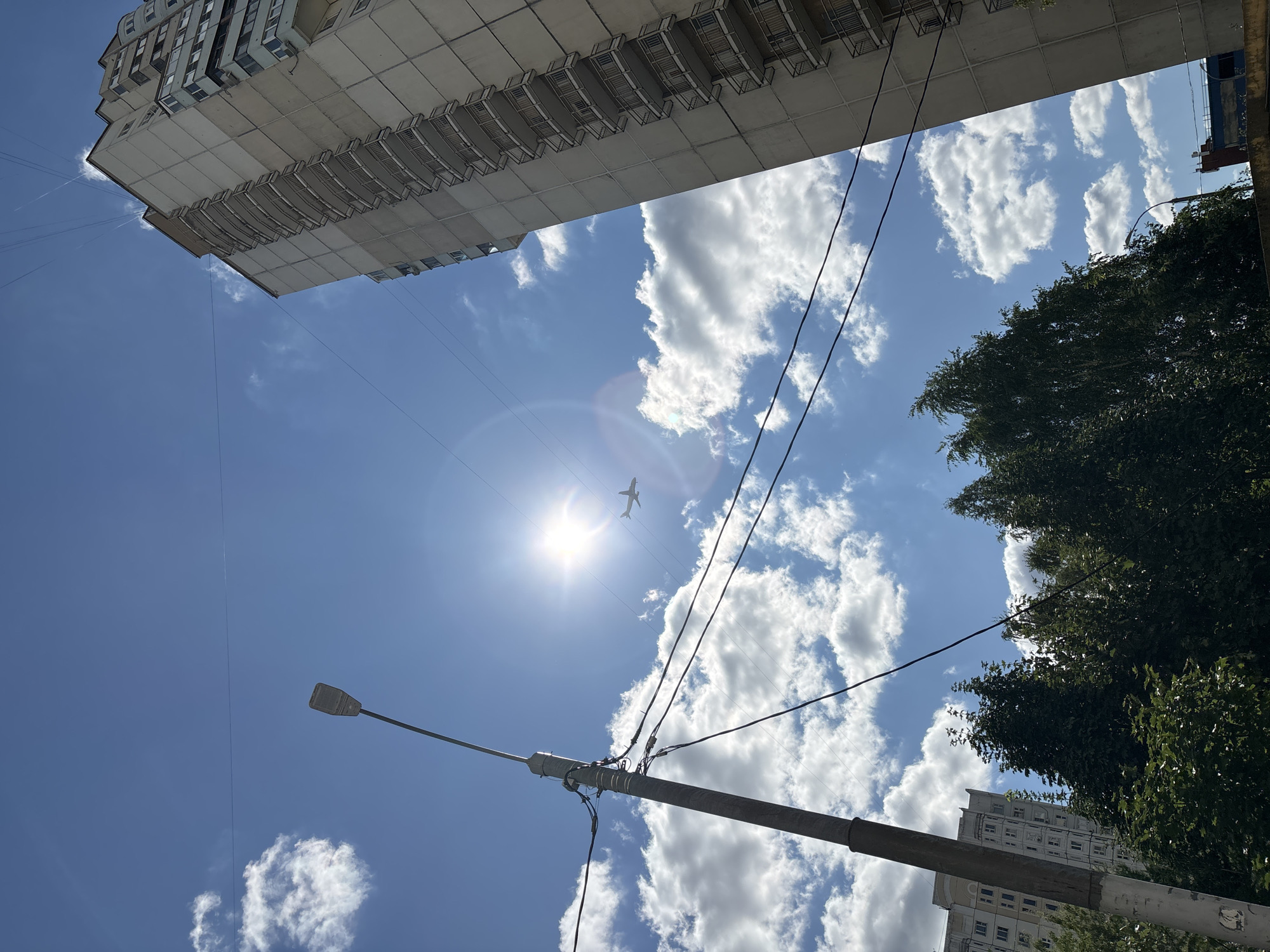 The image depicts a bright, sunny day in a city with a clear blue sky and scattered white clouds. A tall, utilitarian apartment building with multiple balconies dominates the right side of the frame. A lamppost stands on the left, with power lines strung between poles and buildings. In the distance, a small airplane is visible against the sun. The bottom of the frame shows the tops of green trees and other, lower buildings. The location is identified as Odintsovo, Russia.