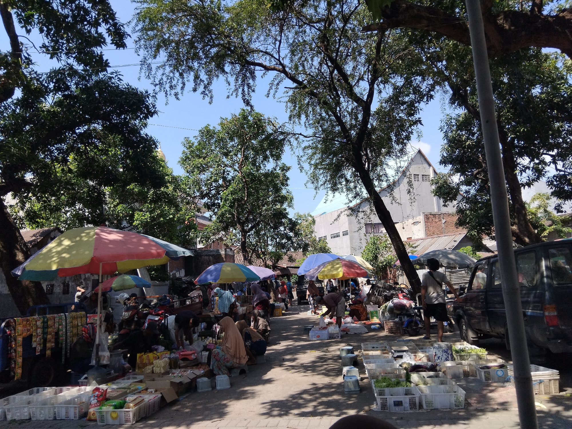 This image captures a bustling outdoor market scene in Kepuh Kiriman, Indonesia. The market appears to be set up along a street or pathway, with numerous vendors displaying their goods. Large, colorful umbrellas provide shade for some stalls, which are laden with various items, including what appear to be packaged snacks, vegetables in white crates, and other assorted merchandise. Several people are visible throughout the market, some attending to stalls, others browsing or interacting with vendors. Motorcycles are parked in the background, suggesting transportation common in the area. Trees line the market, their foliage creating dappled shadows on the ground. Buildings are visible in the background, including a light-colored building with a peaked roof. The overall impression is one of a vibrant local marketplace in operation on a sunny day.