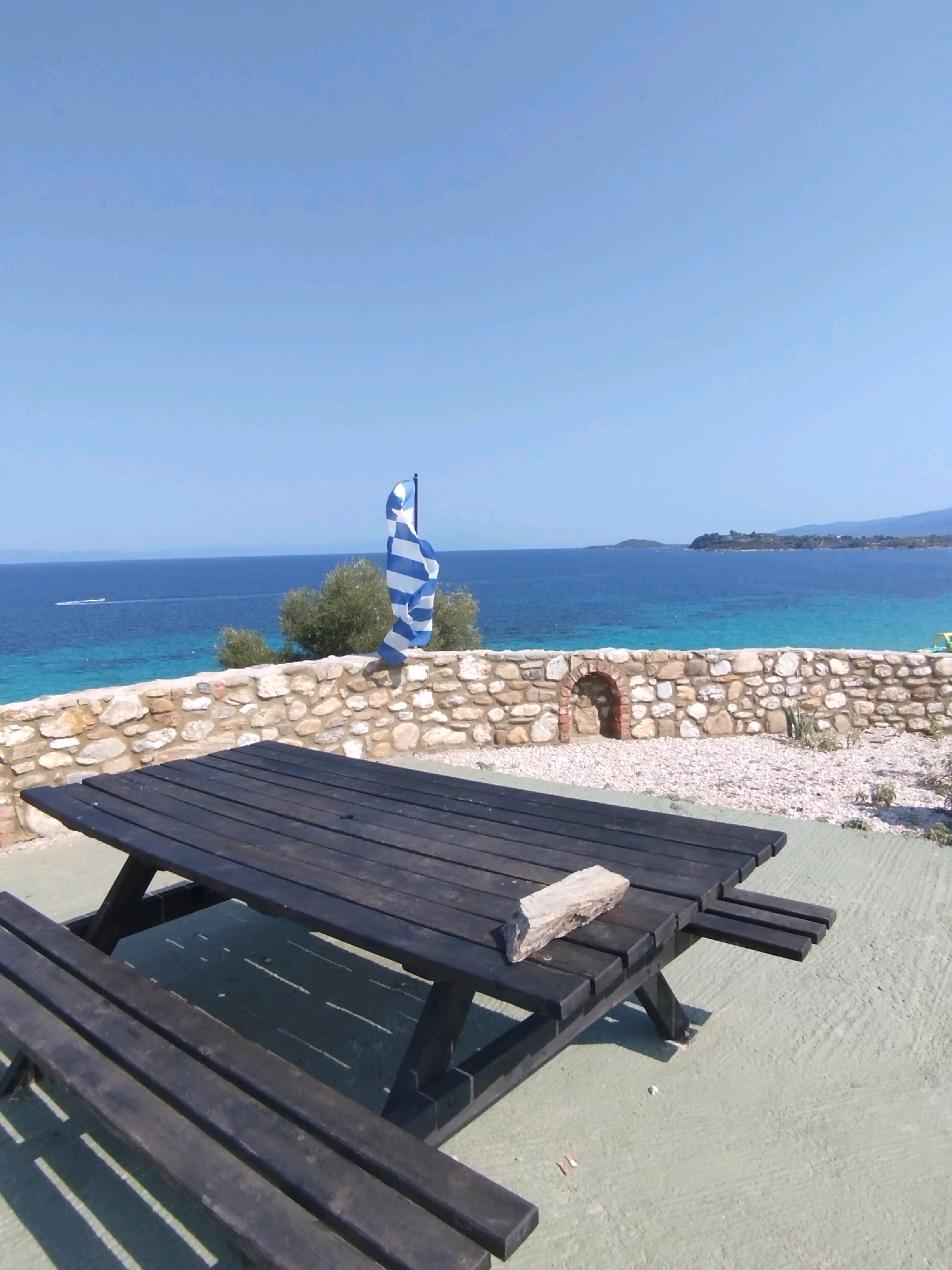 A picnic table and benches made of dark wood are set on a light green paved surface. The table has a small piece of wood resting on its surface. Behind the table, a low stone wall made of various sized rocks forms a barrier. Beyond the wall, the turquoise waters of the sea are visible, with a faint horizon line of land in the distance. A flag with blue and white stripes is planted on the left side of the scene, waving in the wind. The sky is clear and blue. The location appears to be an outdoor area with a view of the sea, possibly in a coastal town or resort. The scene is peaceful and sunny.