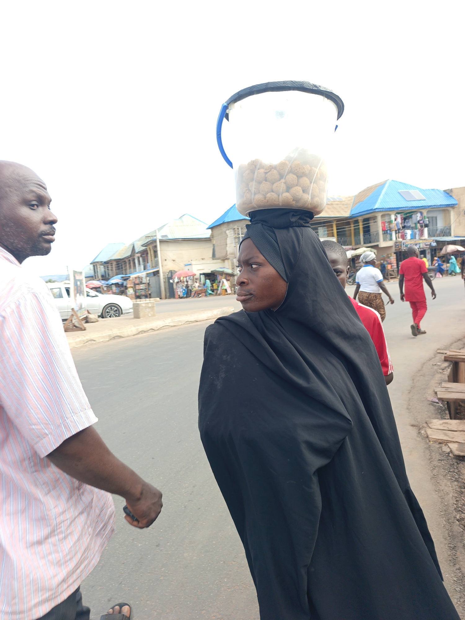 A medium shot depicts a woman in Akwanga, Nigeria, walking on a paved road. She is wearing a long black hijab and balancing a clear plastic bucket filled with round, brown objects on her head. A bald man in a pink striped shirt is walking next to her, looking towards her. In the background, there are buildings with blue roofs, a white car, and other people walking on the street. There are wooden benches along the side of the road. The sky is overcast. The scene suggests a bustling street market or commercial area in Akwanga.