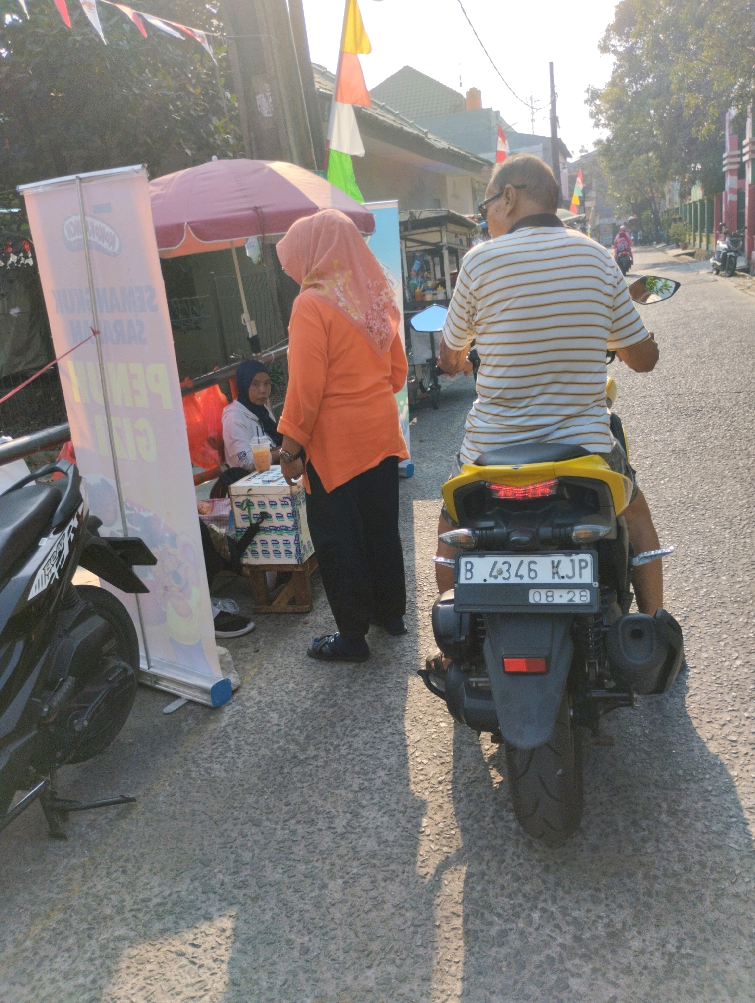 In a city street in Bekasi, Indonesia, a man wearing a striped t-shirt rides a yellow motorcycle. In the background, a woman in an orange shirt and headscarf stands near a stall. A banner with Indonesian text is visible to the left of the stall, and several flags are strung across the street. The scene appears to be a snapshot of daily life in a bustling Indonesian city.
