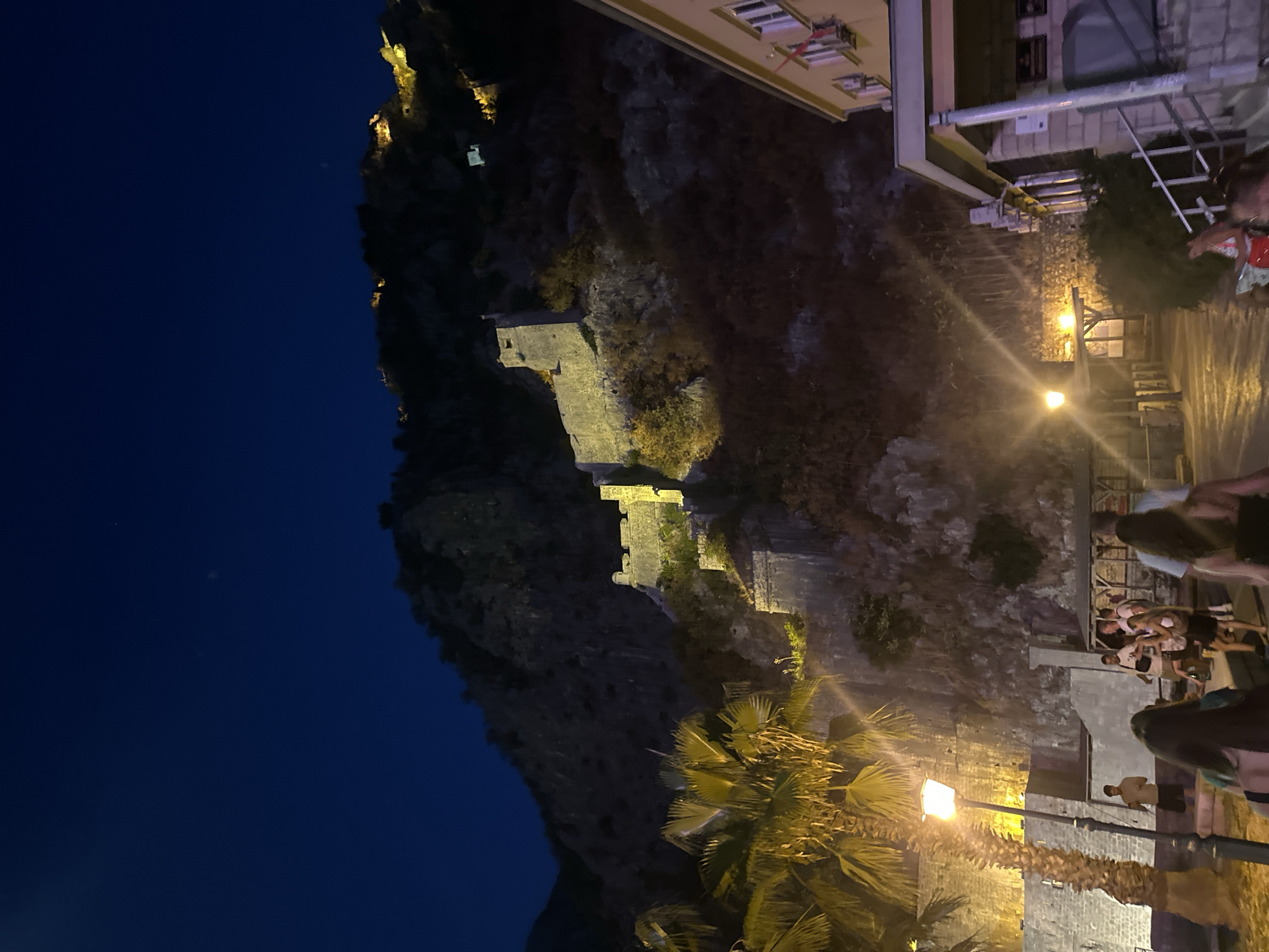 The image depicts a nighttime scene in Kotor, Montenegro, focusing on the ancient fortress walls that ascend a steep mountainside. The fortress, illuminated by artificial lights, stands as a prominent structure against the dark blue night sky. In the foreground, a palm tree with bright yellow fronds is visible, adjacent to a lamppost that casts a warm glow on the scene. A group of people are walking along a paved pathway in front of the fortress, some are conversing. To the right of the image, a building with yellow facade and several windows is present. The overall atmosphere suggests a tourist area at night, with the historical landmark serving as the main visual anchor.