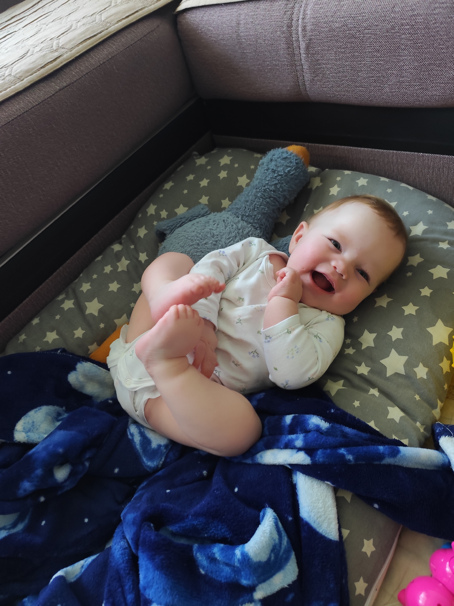 A baby lies on a grey and white star-patterned cushion inside a cushioned area resembling a crib or playpen, partially covered by a blue blanket with white swirls. The baby, wearing a white onesie with small blue floral patterns, is holding their feet in their hands and smiling broadly with an open mouth. A stuffed grey duck toy with an orange beak lies near the baby's head on the cushion. The setting appears to be indoors and the photo does not contain any information indicating the location is in Фоки, Russia.