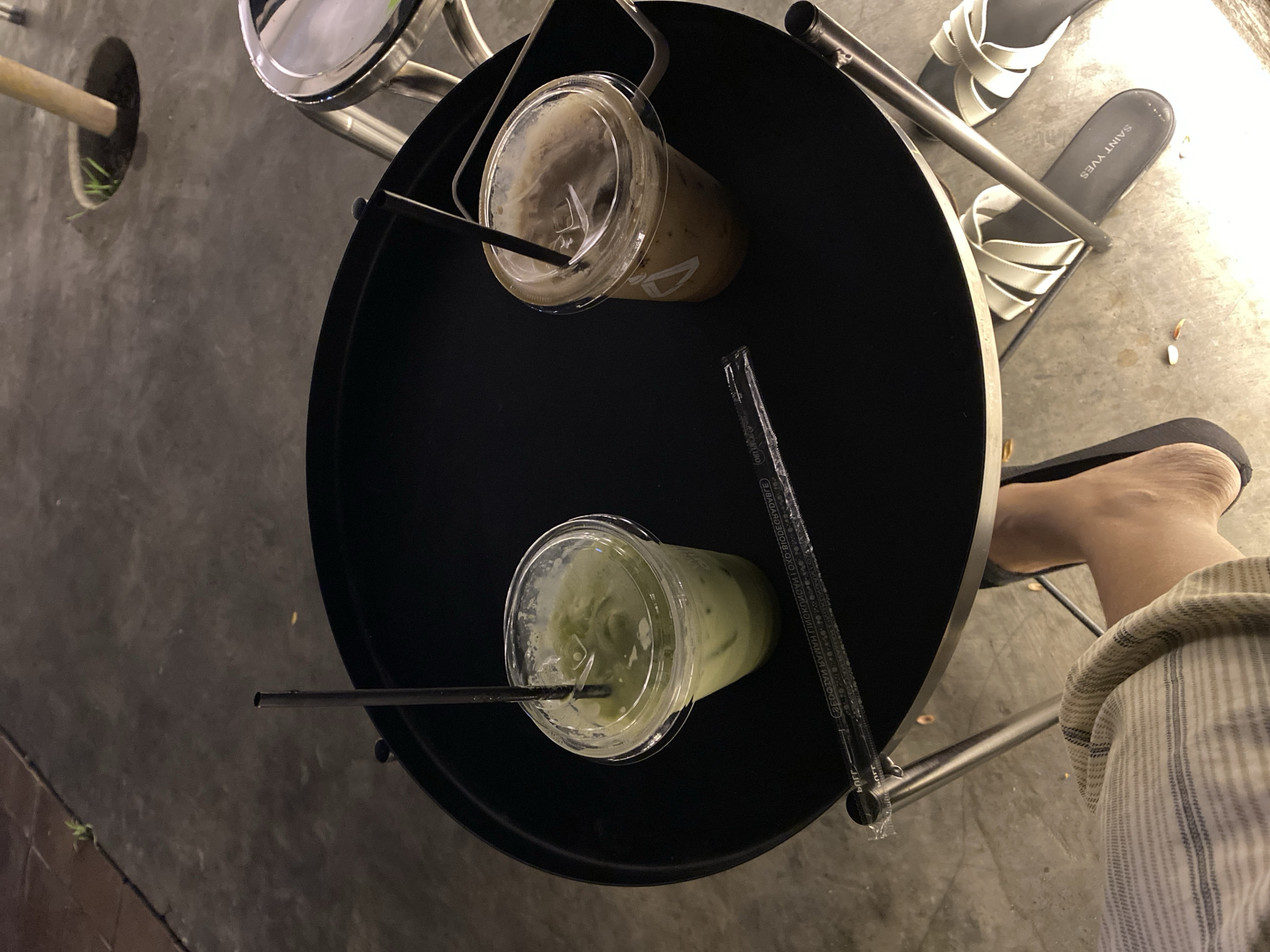 Two iced drinks, one green and one brown, sit on a black circular table. Each drink is in a clear plastic cup with a clear plastic lid and a black straw. A single black straw lies on the table next to the drinks, and a wrapper for straws is also visible. The table has a metal base and is outdoors, with a concrete ground. A person's foot, wearing a sandal, is visible at the bottom of the image. The overall lighting suggests it is nighttime or dusk. The image does not contain enough information to determine the specific location in Madiun, Indonesia.