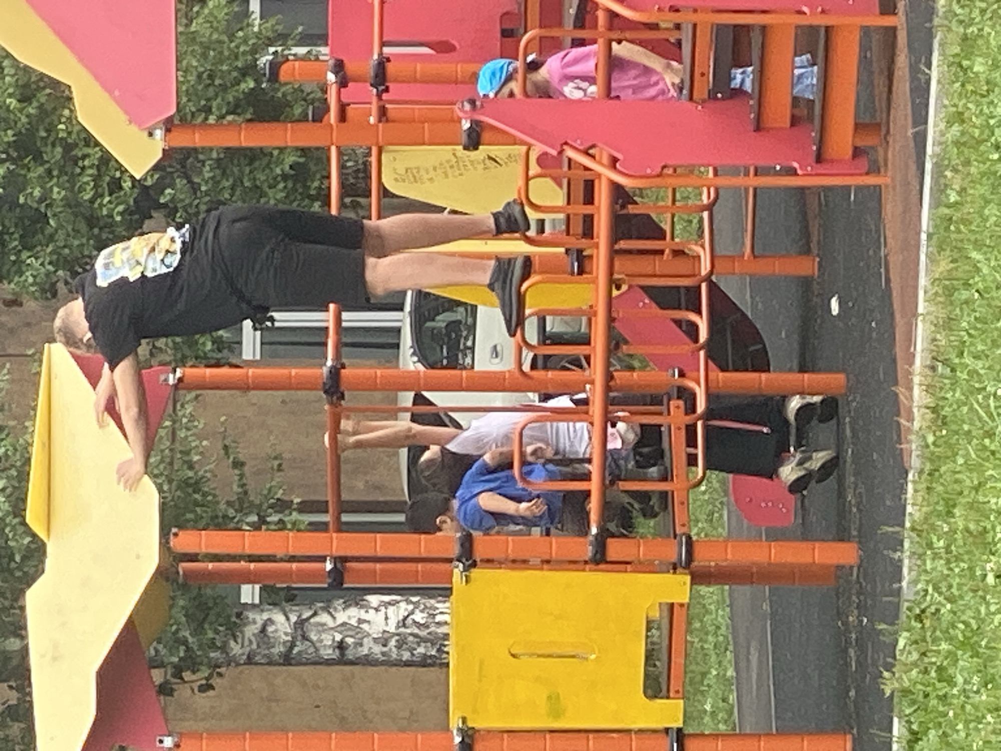 The image depicts a group of people on a playground in Novokuznetsk, Russia. In the foreground, a child in a pink t-shirt and blue headscarf is visible on a set of stairs. To the left, a man in a black t-shirt and shorts is standing on a platform, reaching towards a yellow panel. Below him, a child in a blue t-shirt is also on the playground structure. Further to the right, another child is partially obscured, wearing dark clothing and sneakers. The playground is constructed with orange, yellow, and pink elements, set against a backdrop of green trees and a building. The ground appears to be paved with asphalt.