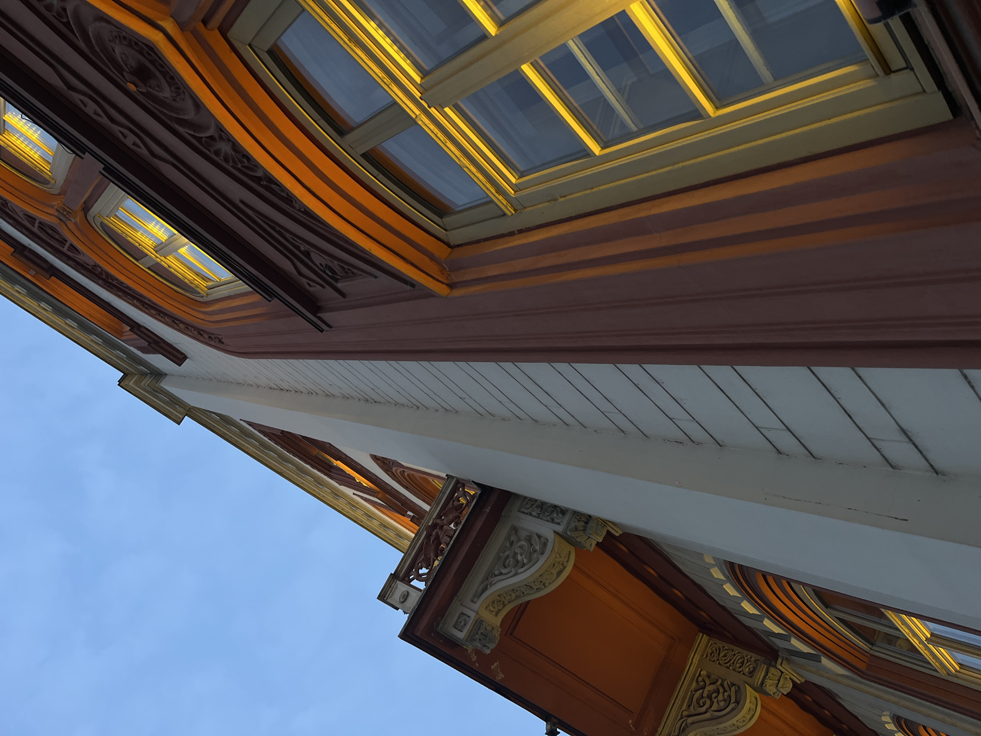 The image presents a low-angle view of an ornate building facade against a clear, pale blue sky, indicating either dawn or dusk. The dominant section of the building features a reddish-brown facade richly decorated with intricate Art Nouveau or Secessionist-style moldings, including stylized organic and geometric patterns. Several windows, some rectangular and others with arched tops, are visible, their light-colored frames encompassing panes that reflect or emit a bright, warm yellow-orange light from the interior. On the left side of the frame, a distinct balcony juts out, showcasing an ornate railing and elaborately carved corbels supporting its structure. The underside of this balcony is painted a vibrant orange. Adjacent to the ornate reddish-brown sections, there are contrasting lighter, possibly off-white or cream-colored vertical wall sections, some exhibiting horizontal relief or siding. No individuals are depicted in the scene. The distinctive architectural style, characterized by its intricate ornamentation, specific color palette, and the elaborate balcony design, identifies the building as **Hotel Moskva**, located on Terazije Square in Belgrade, Serbia.