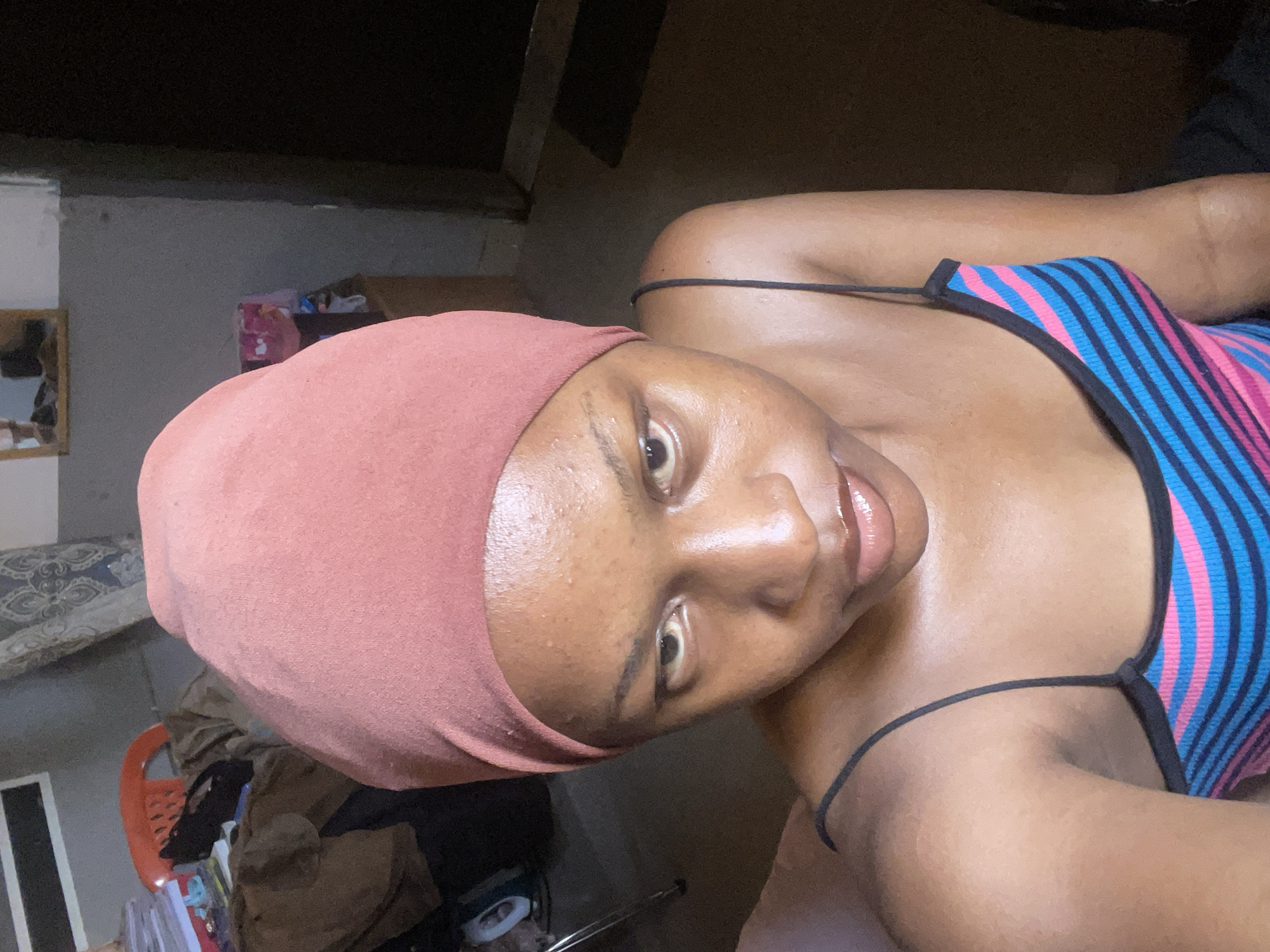 The image features a young woman wearing a dusty rose headscarf and a striped tank top with pink, blue, and black stripes. She is positioned in what appears to be a residential setting, with a dimly lit background that includes furniture and personal items, possibly a bedroom or living area. The environment seems to be indoors, with muted lighting that suggests either early morning or late evening, or simply an interior space with limited natural light. The overall tone of the image is casual and intimate, as it is a selfie. The woman's expression is direct and engaging, with a slight smile and her gaze focused on the camera. Her skin appears to be glowing, possibly due to makeup or natural lighting. The focus is primarily on the woman herself, with the background elements slightly out of focus, drawing attention to her features and attire. There are no visible signs of text in the image. The implied context of Jos, Nigeria, places the scene within a specific geographical and cultural environment, though the visual cues themselves do not explicitly reveal this location.
