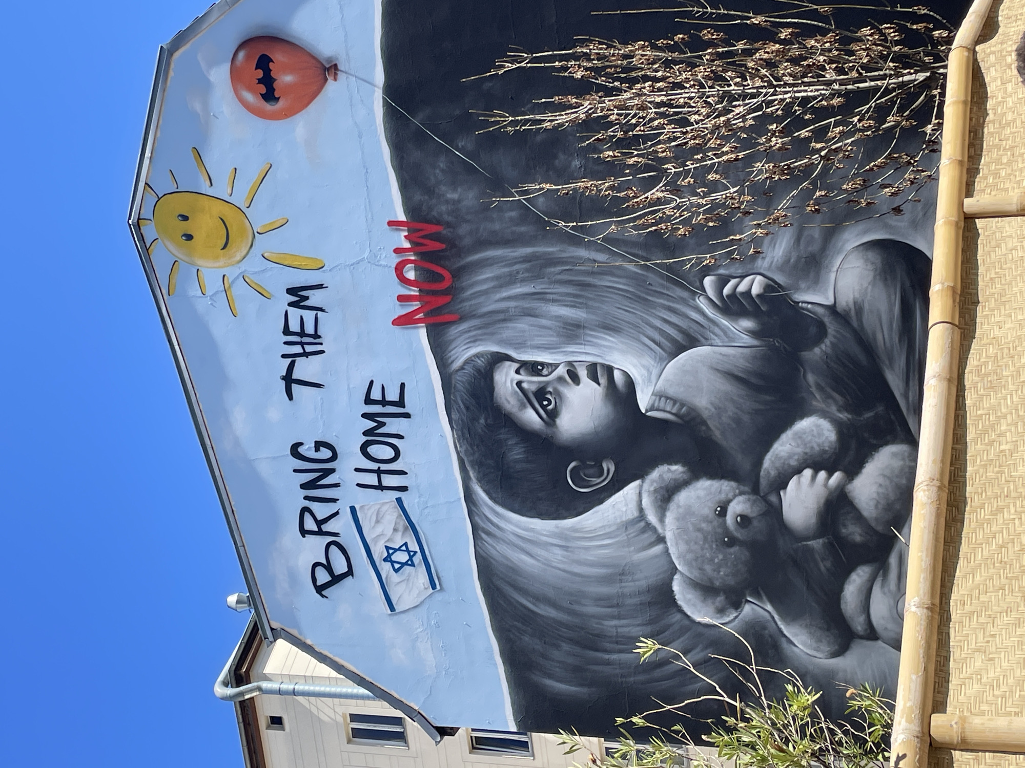 A mural is depicted on the side of a building in Berlin, Germany, under a clear, bright blue sky, suggesting daytime. The mural is divided conceptually into two main parts. The left side features a light blue background with a cheerful yellow sun in the upper left corner and an orange balloon with a black Batman symbol floating near it. Below the sun, the phrase "BRING THEM HOME" is written vertically in black, with a small depiction of an Israeli flag positioned beneath it.
The right side of the mural is predominantly grayscale, portraying a child looking upwards with a distressed expression, clutching a teddy bear. The word "NOW" is painted prominently in red, overlapping the grayscale depiction of the child. Bare tree branches extend into the frame from the right, partially obscuring the upper right portion of the mural. The overall image conveys a strong emotional message, advocating for the return of individuals, likely referring to hostages, emphasized by the child's somber depiction and the urgent red "NOW."