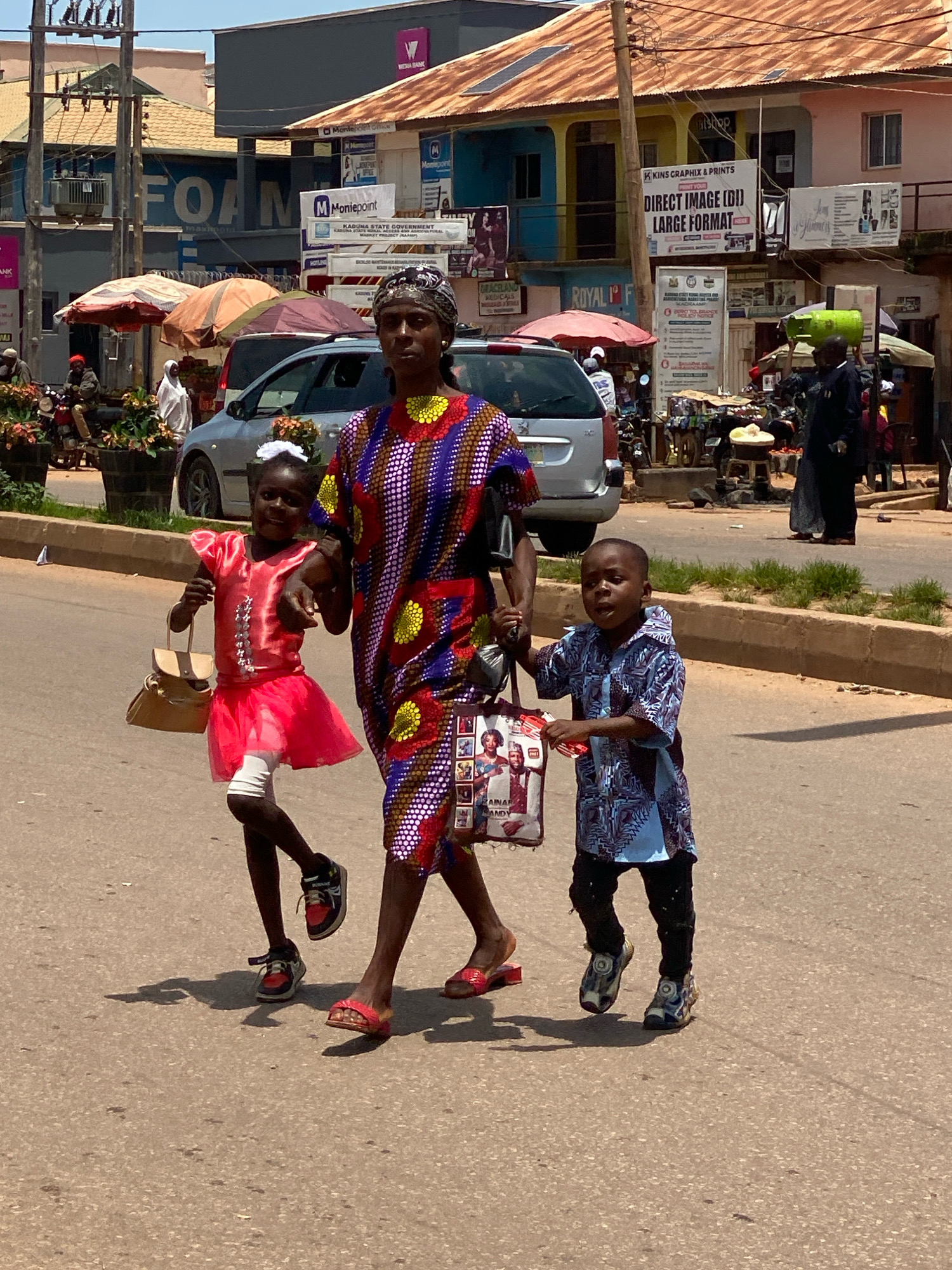 In Sabon Tasha, Nigeria, under a bright, sunny sky, a group of people navigate a busy street. A woman, dressed in a vibrant, multi-colored patterned dress and headwrap, walks purposefully, holding onto a shopping bag. She is accompanied by two children. A young girl, wearing a bright pink dress and carrying a tan handbag, walks beside her. A young boy in a blue patterned shirt and black pants, also holding a shopping bag, walks on the other side. They are walking on a paved road with a concrete curb lined with grass. In the background, shops with various signs, motorcycles, parked cars, and street vendors are visible. The scene captures a moment of everyday life in a bustling urban environment.