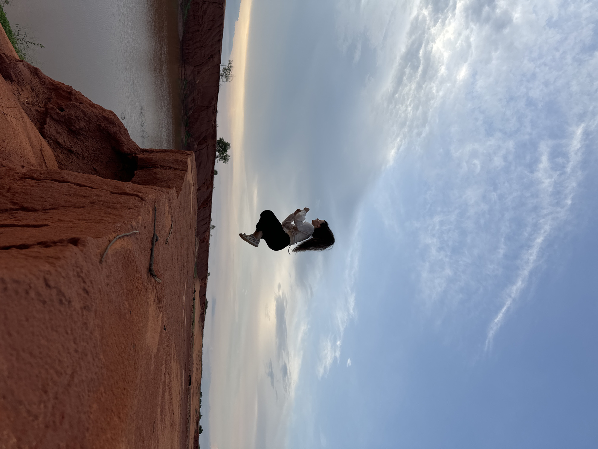 A young woman is captured mid-air, appearing to jump or float, against a striking natural landscape. She has long, dark hair and is dressed in a white long-sleeved shirt, black pants, and light-colored sneakers. Her body is slightly curled, conveying a sense of weightlessness and movement.
The scene below her features steep, rugged reddish-brown cliffs or sand dunes, characterized by their earthy tones and distinct textures. These formations border a calm body of water, likely a river or lake, visible along the lower and left side of the frame. Sparse green vegetation, including small trees, dots the distant banks.
The expansive sky above her dominates the upper and right portions of the image. It transitions from a soft blue at the top to a lighter, warmer, possibly golden hue near the horizon, suggesting either early morning or late afternoon light. Scattered, wispy white clouds add texture to the sky. The overall impression is one of freedom, serenity, and a connection with nature. This landscape, with its prominent red earth formations and water body, is consistent with areas like the Fairy Stream or Red Sand Dunes in Mui Ne, Vietnam. There is no visible text.