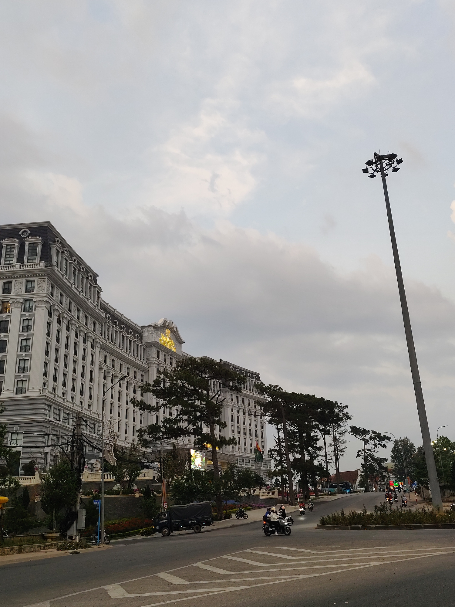 The image shows the Muong Thanh Da Lat Hotel in Da Lat, Vietnam. The multi-story white hotel building is on the left, with trees obscuring the lower levels. A tall light pole is on the right. The sky is overcast. A street intersection in the foreground has a few vehicles, including motorcycles and a truck, and a few pedestrians.