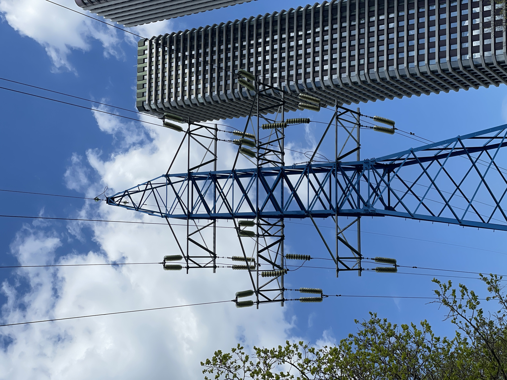 The image depicts an upward view of a blue metal electric power transmission tower against a blue sky with scattered white clouds. Above the tower is a modern building with a repetitive pattern of windows and horizontal architectural details. The tower is strung with high-tension power lines supported by insulators. The base of the image shows the green foliage of trees. The image was taken in the city of посёлок совхоза "Останкино", Russia.
