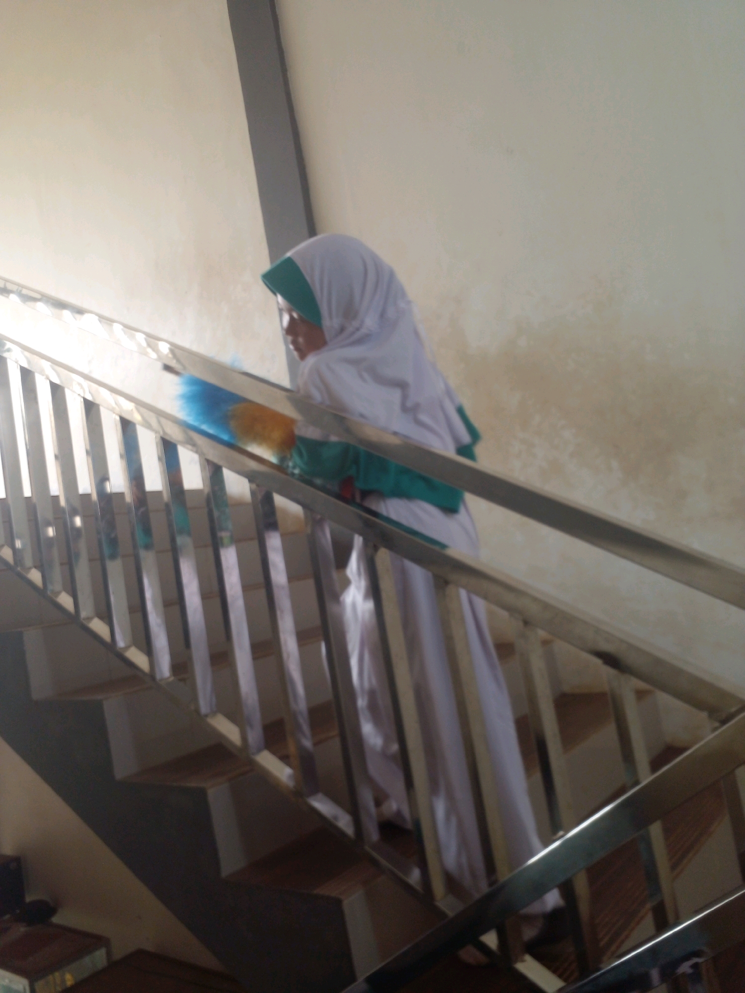 A person wearing a white hijab and a green trim is on a staircase, holding a blue and yellow duster. The staircase has wooden steps and a metal railing. The walls are light-colored, and there is natural light coming from the left. The scene appears to be indoors.