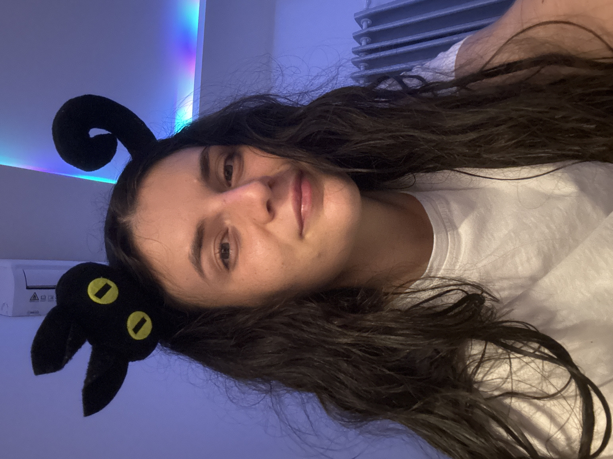 A woman is wearing a black cat-ear headband. She is lying on a white surface with dark, wavy hair spread around her. In the background, there is a radiator, a white air conditioner, and a color-changing LED strip. The image does not contain information to confirm the location is Mũi Né, Vietnam.