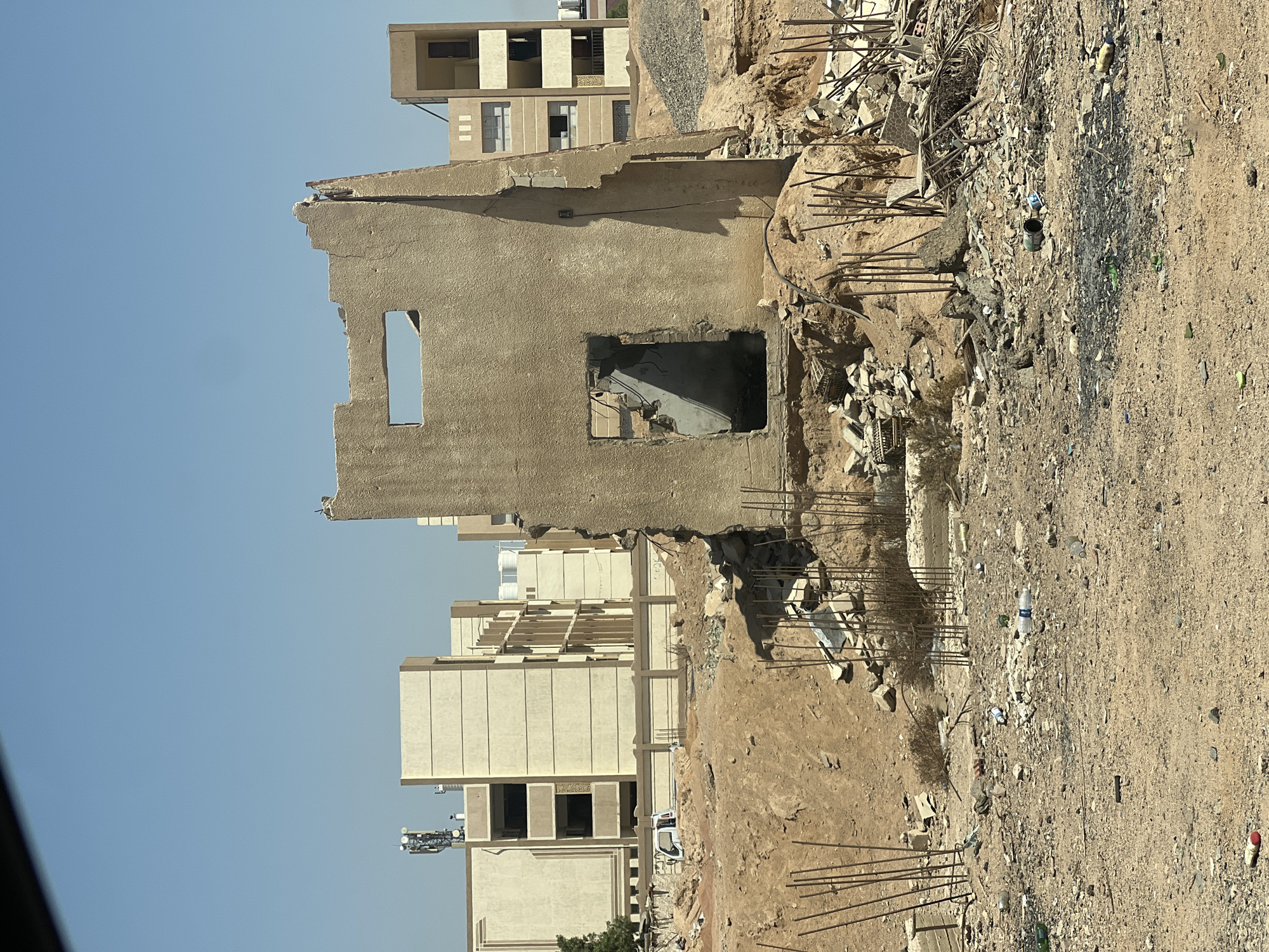 The image depicts a desolate construction site in Sharm El Sheikh, Egypt, characterized by the remnants of a partially demolished building. In the foreground, the ground is littered with debris, including rebar, broken concrete, and scattered plastic bottles. A partially standing, rough concrete structure with a rectangular opening and a dark interior dominates the center of the frame, showing signs of significant structural damage. Behind this ruined structure, several modern apartment buildings stand intact, their beige facades contrasting sharply with the surrounding desolation. A low concrete fence separates the construction site from the completed buildings. A white pickup truck is visible behind the fence. The sky above is a clear, pale blue. The overall impression is one of ongoing or halted construction and demolition in an arid, urban environment.