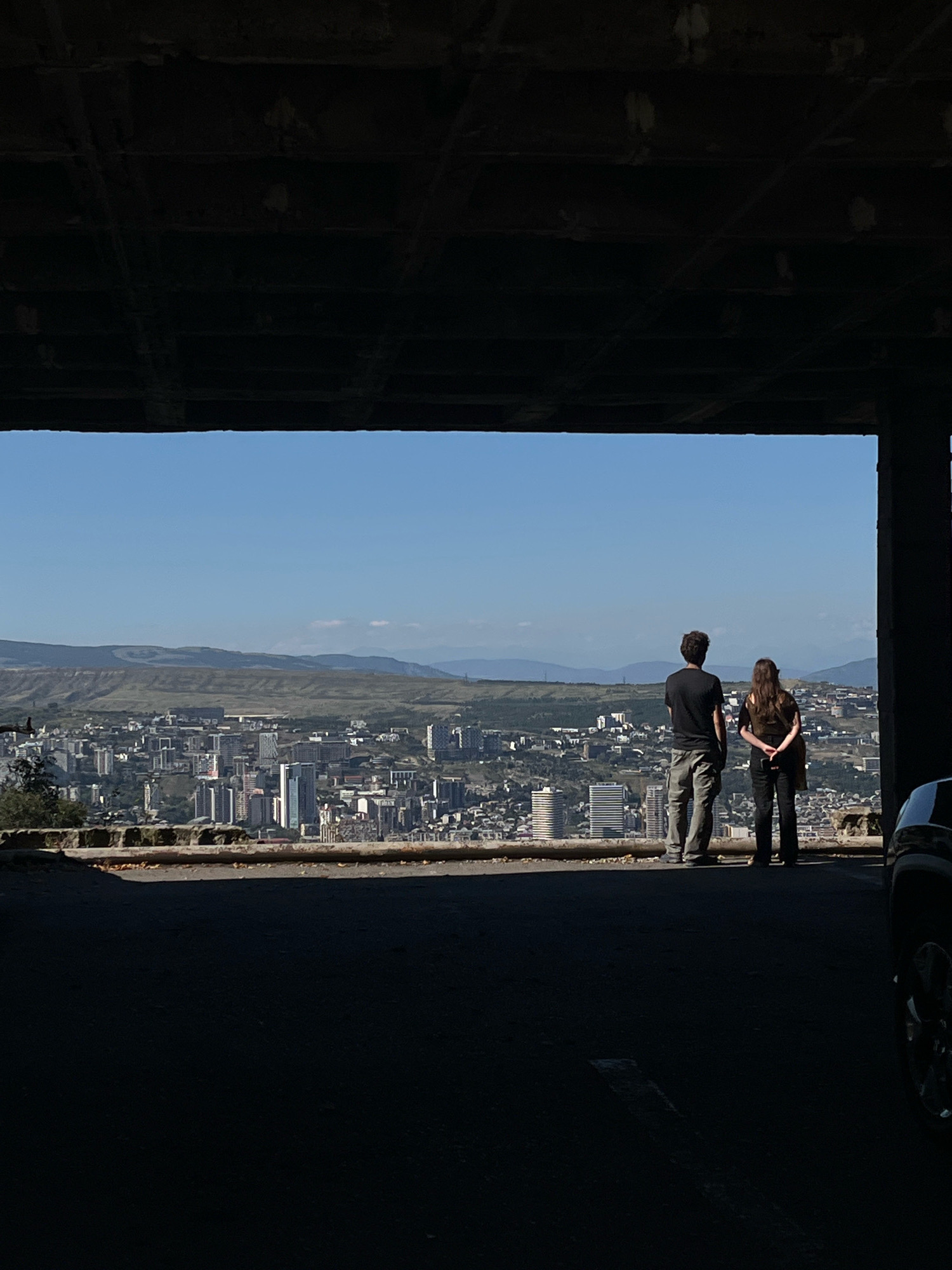The image shows a panoramic view of Tbilisi, Georgia, seen from a high vantage point, likely from an elevated highway or an unfinished structure, as indicated by the dark, shadowed foreground and ceiling. Two individuals, a man and a woman, stand on a paved surface, looking out at the sprawling cityscape. The man is wearing a black t-shirt and light-colored cargo pants, while the woman is dressed in a dark top and pants, carrying a bag over her shoulder.
The city of Tbilisi is spread out below, characterized by a dense collection of buildings with varied architectural styles, including modern high-rises and older structures. The landscape beyond the city is marked by rolling hills and mountains under a clear blue sky. The foreground is dominated by shadows, suggesting the viewpoint is from within a covered area, possibly an underpass or an abandoned building. A portion of a dark-colored vehicle is visible on the far right of the frame. The overall impression is one of urban exploration or enjoying a scenic overlook.