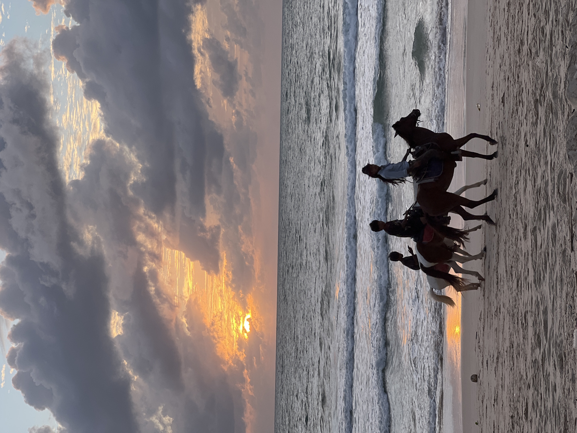 In the city of Choeng Thale, Thailand, a tranquil beach scene unfolds at sunset. The sky is a dramatic canvas of dark, textured clouds interspersed with bright orange and yellow hues from the setting sun. The sun, partially obscured by clouds, casts a warm glow on the horizon, reflecting a shimmering path across the ocean's surface. The ocean itself is moderately active, with several waves breaking near the shore, creating white foam that washes onto the sandy beach.
On the beach, three individuals are riding horses. The person in the foreground on the right is riding a dark brown horse and appears to be wearing a hat. To the left and slightly behind are two figures on a lighter-colored horse. The rider in the middle is a child, and the person behind them is an older child or adolescent. The horses are moving along the wet sand, their hooves leaving imprints. The overall atmosphere is peaceful and serene, with the natural beauty of the sunset over the sea creating a picturesque setting.