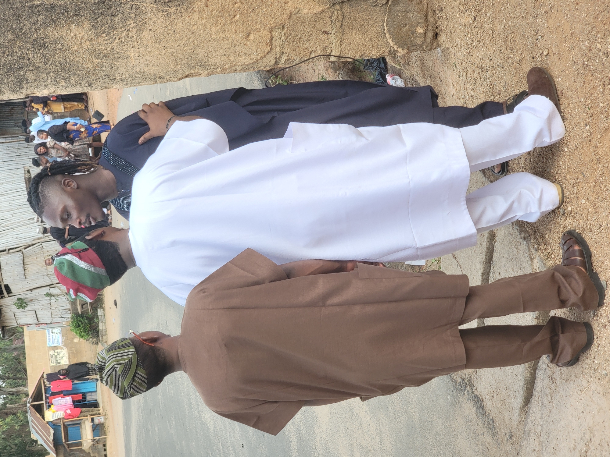 The image shows three men standing in a row on a paved surface that appears to be a street. The men are likely in Jos, Nigeria, suggested by the traditional clothing. The first man wears a brown robe-like garment over matching trousers and brown sandals, and a striped cap. The second man is dressed in a white robe and trousers. The third man wears a dark blue robe over white trousers and brown shoes, his hair is styled in dreadlocks. A group of people is visible in the background, along with buildings and a street-side shop with stacked colorful containers. The surface around the men appears to be a mix of pavement and dirt.