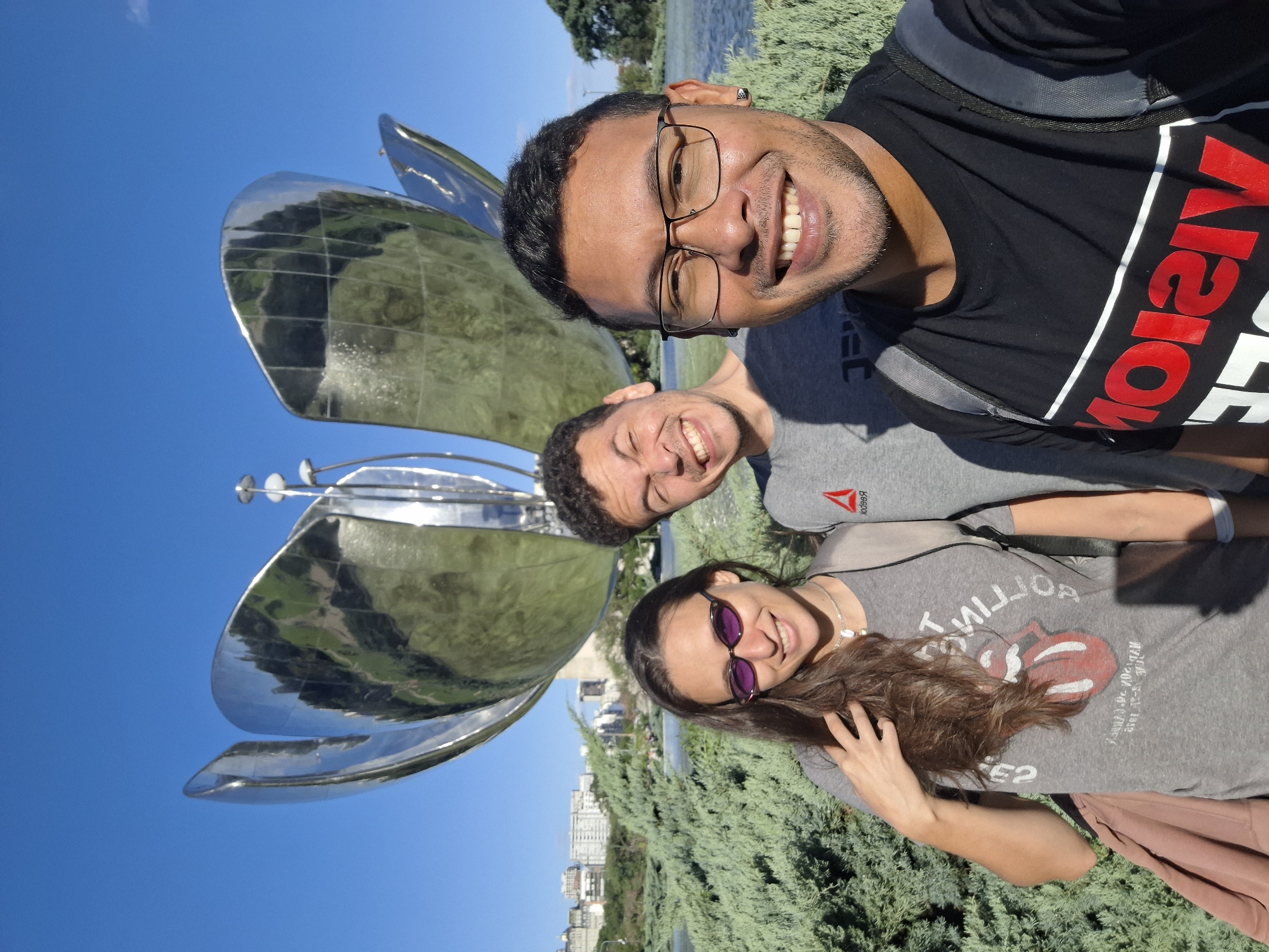 Three individuals are posing for a selfie in front of the Floralis Genérica, a large metal flower sculpture located in Buenos Aires, Argentina. The sculpture is composed of reflective metal petals, with the blue sky reflected in the metal. The foreground features green vegetation and the cityscape of Buenos Aires is visible in the background. The individuals are smiling. The person in the front is wearing glasses and a black T-shirt with a red logo, and a gray backpack. The person behind is wearing a gray T-shirt with a Reebok logo. The woman is wearing sunglasses and a Rolling Stones T-shirt.