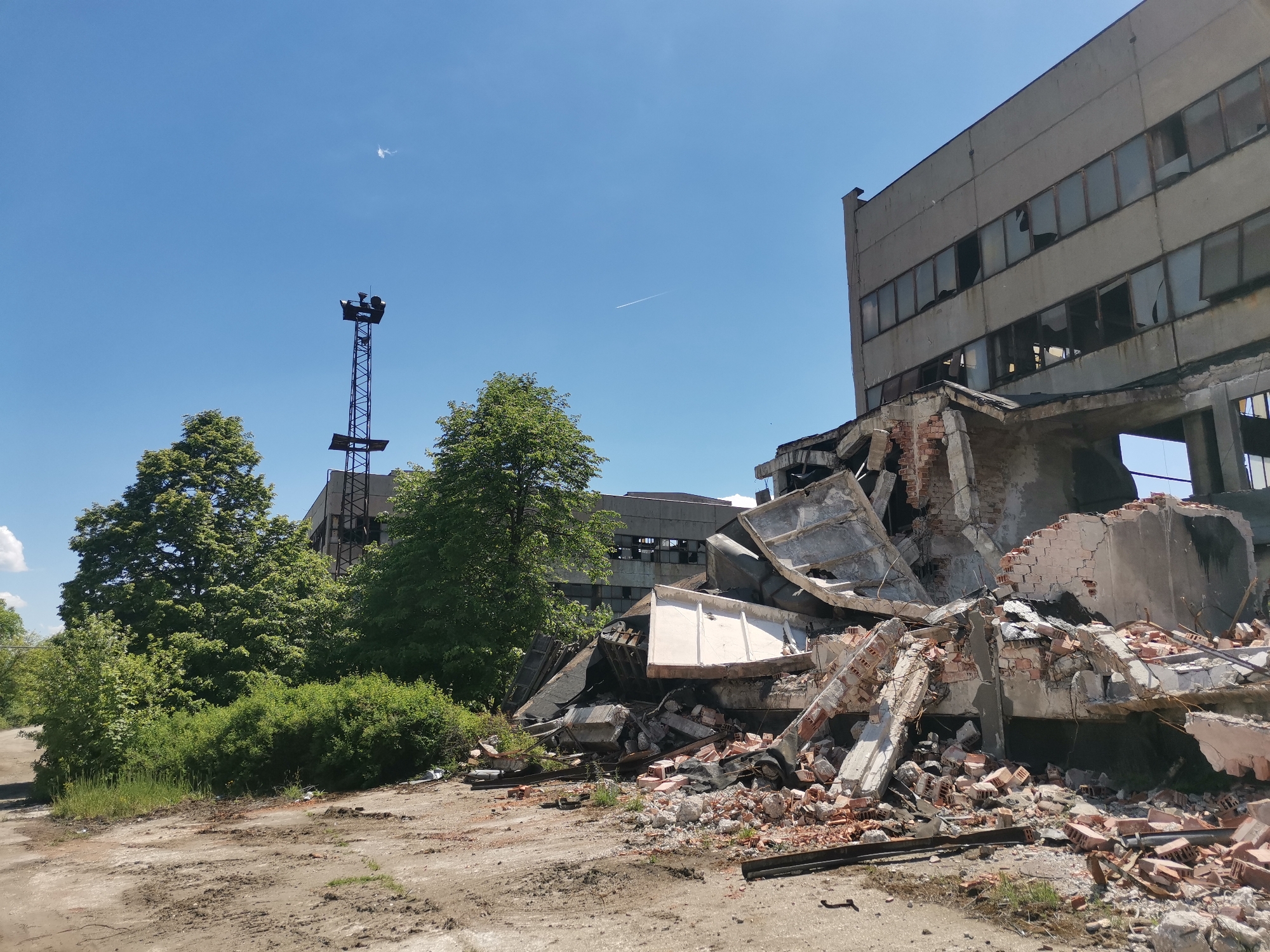 The image depicts a scene of demolition and decay. A partially destroyed concrete building with shattered windows dominates the right side of the frame, with a heap of crumbled brick and concrete debris at its base. In the background, a slender metal tower stands adjacent to greenery and other buildings. The ground is a mix of bare earth and cracked pavement. The sky is clear and blue. There is no evidence to suggest this is Zhivkovo, Bulgaria. There are no people present in the image.