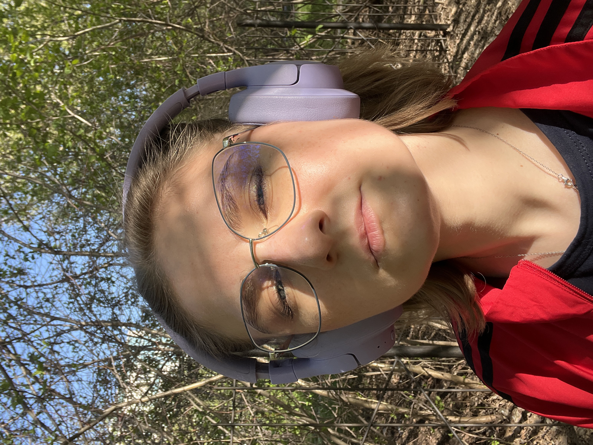A young person with closed eyes, wearing lavender headphones and round metal-framed glasses, is outdoors. They are wearing a red and black jacket and a silver necklace. The background consists of trees, branches, and patches of blue sky. There is no indication of Novosibirsk, Russia.