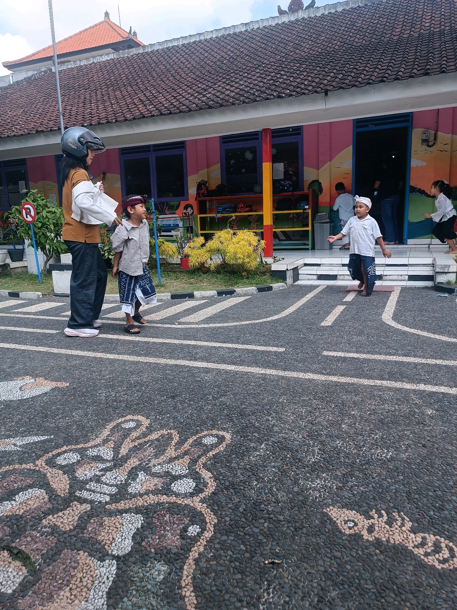 This is a medium-shot, eye-level, outdoor photograph taken in front of a colorful school building. The building has a tiled roof and a mural painted on its facade. In the foreground, a boy wearing a white shirt and a sarong is walking on a paved area with a road design. The pavement is adorned with a mosaic of stones forming abstract shapes.
To the left, a woman wearing a brown shirt and dark pants, with a helmet on her head, is standing next to another boy who is also wearing a white shirt and a patterned sarong. They appear to be observing the boy in the middle. Behind them, the entrance of the school building is visible, with other children and adults inside. To the right, another child, a girl in a white top and dark pants, is running.
The environment suggests a schoolyard or an outdoor activity area. The presence of children in traditional attire and the style of the building might indicate a cultural or educational institution in Indonesia, possibly in Tabanan, Bali, given the architectural elements and the attire. The sky is overcast, suggesting a diffused natural light.
