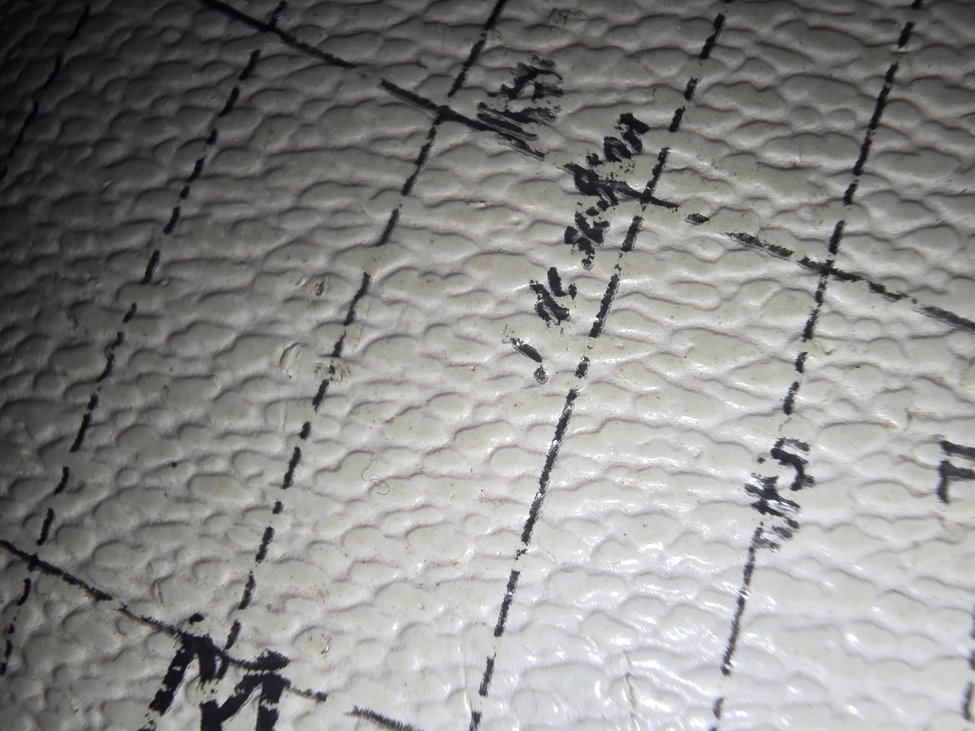 The image is a close-up, macro photograph of a white, textured surface with black markings and lines. The texture appears to be from a ball, possibly a basketball or volleyball, due to the dimpled pattern. There are dashed black lines, solid black lines, and what appears to be handwriting or smudged text in black ink. The lighting is uneven, creating highlights and shadows that emphasize the texture of the surface. The image lacks any discernible context or features that would allow for identification of a specific location within the city of Ulan-Ude, Russia, or any other urban environment. There are no people, animals, or any other identifying elements present.
