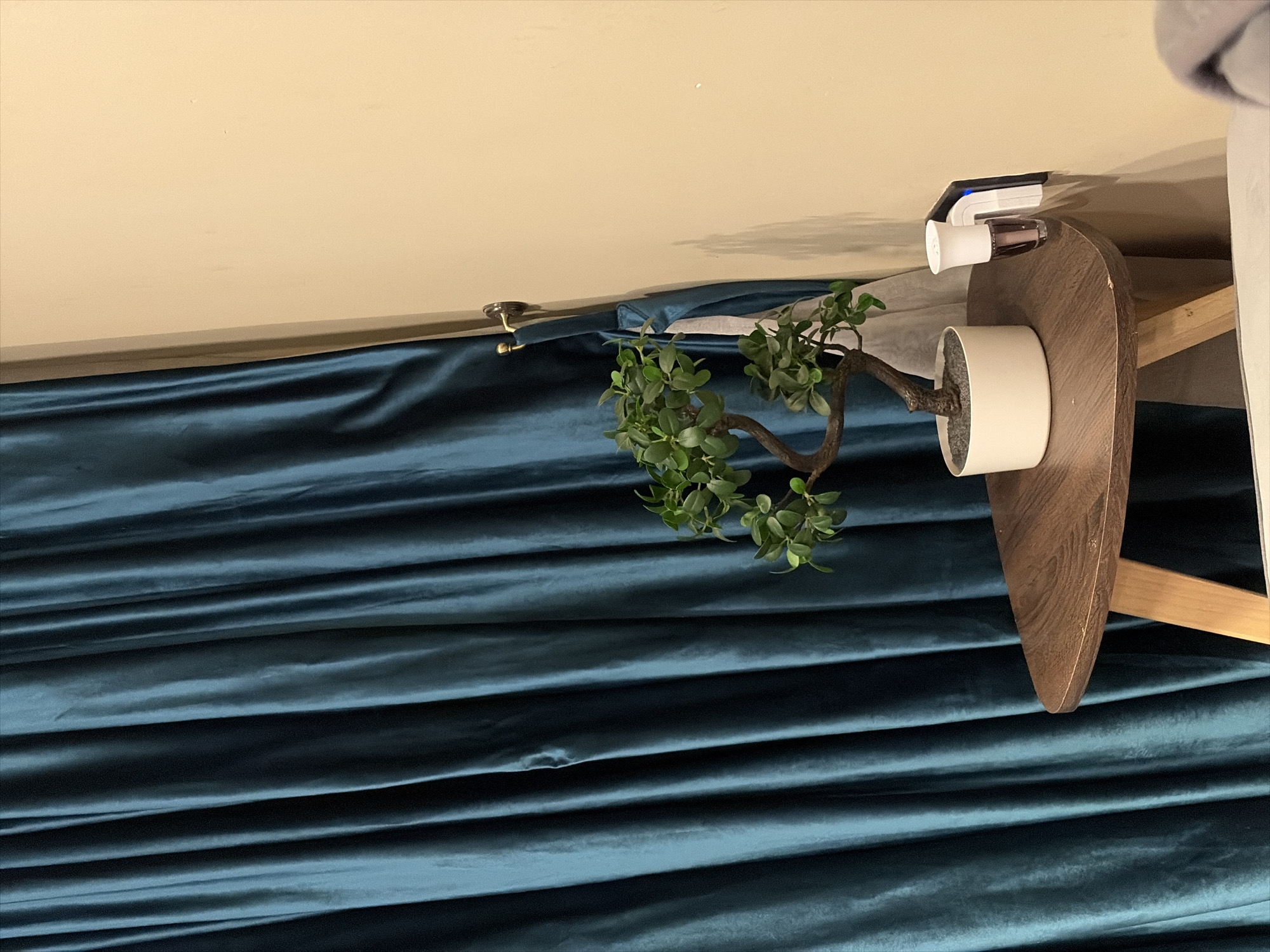 An indoor scene, possibly within a home in Abuja, Nigeria, features a meticulously arranged setting with a focus on textures and tones. The left side of the frame is dominated by a plain, light beige or cream-colored wall, creating a clean backdrop. To the right, a rich, deep teal or dark blue curtain, likely made of velvet or a similar plush fabric, hangs in thick, horizontal folds, adding depth and a sense of luxury to the space.
In the foreground, a small, dark brown wooden side table with a distinctively rounded, oval-shaped top is visible. Resting on this table is a small, potted ornamental plant, resembling a faux bonsai, characterized by its vibrant green leaves and a gnarled, brown stem, contained within a simple white cylindrical pot.
Above the table, on the wall, a small white and silver/grey electronic device with a distinct blue indicator light is plugged into a wall socket, suggesting a modern utility like a smart plug or a small charger. Further to the left on the wall, a brass-colored curtain tie-back or hook can be seen, indicating where the curtain might be gathered. The overall lighting is soft and ambient, creating a calm and inviting atmosphere. No people are visible, nor is there any discernible text. The original media file is rotated 90 degrees clockwise, with this description assuming a corrected, upright orientation of the room.