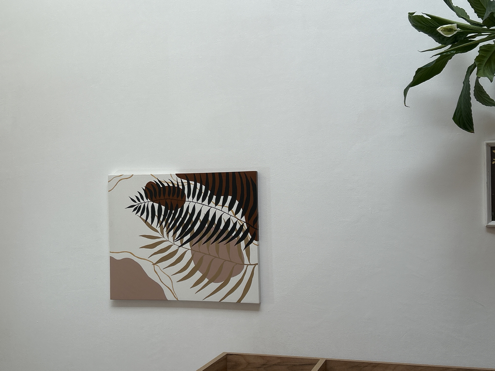 A close-up shot of a white wall, showcasing a square art piece with a stylized design of botanical elements in brown, black, and gold tones, and a houseplant with green leaves at the upper right. The lower part of a wooden structure is visible in the bottom of the frame. The location is implied to be in Chaikovsky, Russia, based on the prompt's instructions. No people are visible.