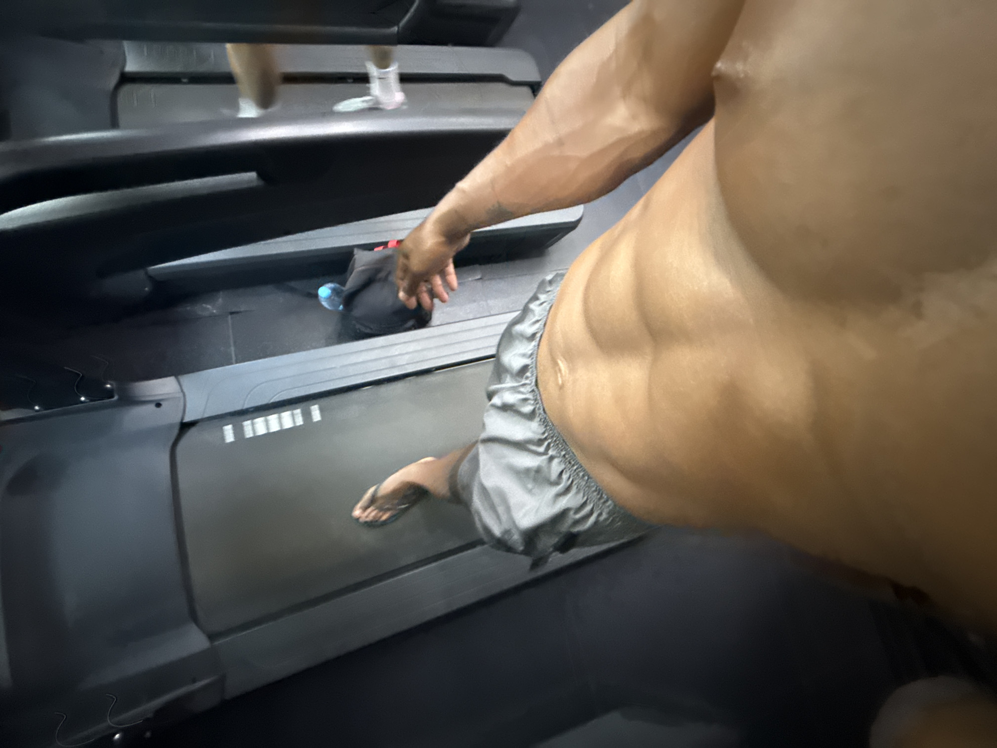 A close-up shot depicts a muscular, shirtless person standing on a treadmill. They are wearing gray shorts. Their right foot rests on the moving belt of the machine. The individual’s left hand is near a black bag and a clear water bottle, which are positioned on the stationary part of the treadmill. A second person's legs are visible on the treadmill ahead, indicating someone else is also using the machine. The location appears to be a gym with black rubber flooring. The city is Unknown, Unknown.