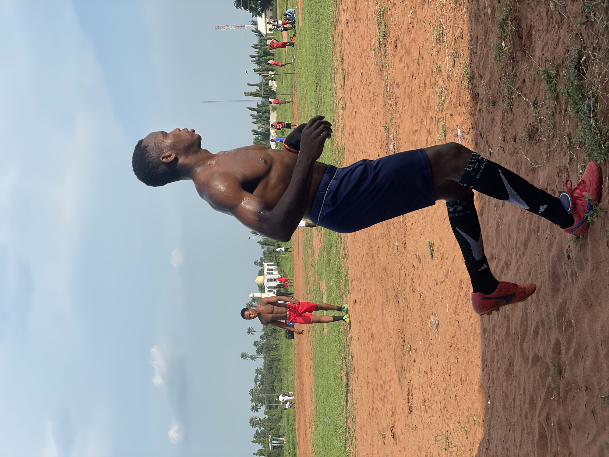 The image displays a dark-skinned man engaged in physical activity on an outdoor sports field under bright, clear daylight in Ogbogbo, Nigeria. The main subject, positioned prominently, is shirtless, revealing a muscular and glistening physique, indicating he is sweaty. He wears dark blue athletic shorts, black and white athletic socks (one clearly showing "Adidas" text and a Nike-like swoosh), and red sports shoes, likely cleats. His head is tilted upwards and slightly backward, with his eyes looking towards the sky, suggesting a moment of exertion, deep breath, or focus during training.
The setting is an open field, characteristic of a football (soccer) pitch, divided into two primary surfaces: a reddish-brown dirt or sand area in the immediate foreground and a verdant green grass section extending into the mid-ground. In the distance, several other individuals are visible on the grass field, sparsely distributed, with one shirtless man in red shorts standing out more clearly.
The background features numerous palm trees and other dense foliage, contributing to the tropical or subtropical feel of the location. A distinctive white building, possibly a mosque or a large house with a dome-like structure, and a tall communication mast are also visible under the expansive blue sky. The bright sun casts strong shadows, indicating a time of day between late morning and late afternoon. The overall atmosphere suggests a warm, sunny day ideal for outdoor sports.