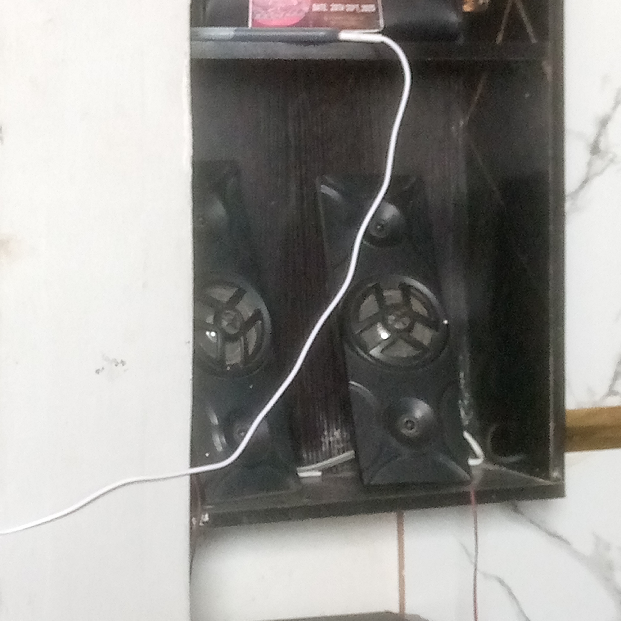 The image shows a shelf with two black speakers. A white cable is draped across the speakers and shelf. The background is a dark, textured surface. The left side of the image shows a white surface, possibly a wall or cabinet.