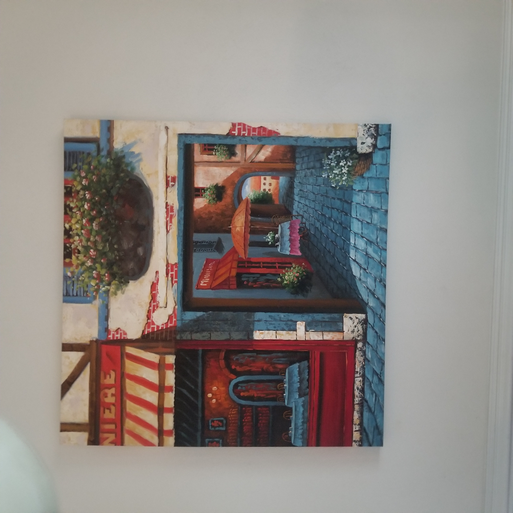 This is a photograph of a painting hanging on a white wall. The painting depicts a quaint cobblestone alleyway in what appears to be a European village. On the left, there is a storefront with a red awning and the word "NIERE" visible above it. Inside the storefront, tables are set for dining.
The alleyway opens up to reveal another shop with an orange awning and a small table with an umbrella. Beyond this, the alley curves and leads to a view of distant buildings under a warm, golden sky. The painting is rendered in a style that suggests oil on canvas, with visible brushstrokes and a focus on light and shadow. There are no people depicted in the painting. The image does not contain any information that would suggest it is located in Virginia Beach, United States of America.