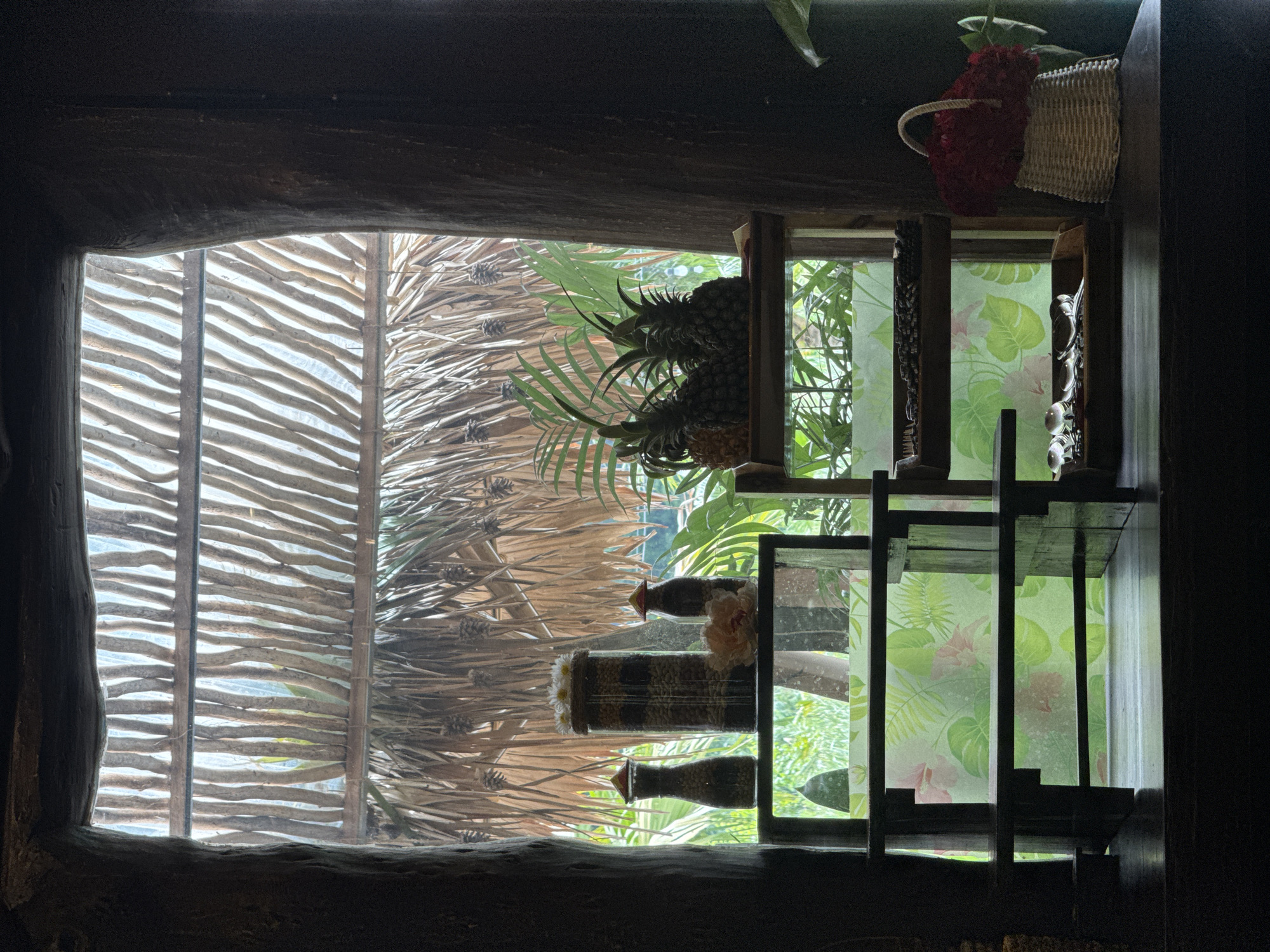 The image depicts an indoor scene with a view through a window to an outdoor tropical environment. The window frame is constructed from rough, dark wood, resembling tree trunks. Through the window, a thatched roof and lush green foliage are visible.
Inside, on a dark wooden surface, are several decorative items. On the left, a tiered wooden shelf holds three containers. The tallest, in the center, is a glass jar filled with what appears to be layered nuts or seeds, topped with small white flowers. Flanking it are two smaller, dark glass bottles with conical lids, also containing visible granular contents. A pale pink artificial flower rests on the shelf.
To the right of the first shelf, a second, lower wooden shelf displays three pineapples. Below this, another shelf has a tray containing coffee beans and a second tray with several silver spoons. To the far right, a white woven basket overflows with a dense arrangement of red flowers.
The background seen through the window shows a vibrant green garden with tropical plants. The overall ambiance suggests a serene, possibly resort or restaurant setting in a warm climate. The location provided, Nam Hoa Lư Ward, Vietnam, aligns with this tropical aesthetic.