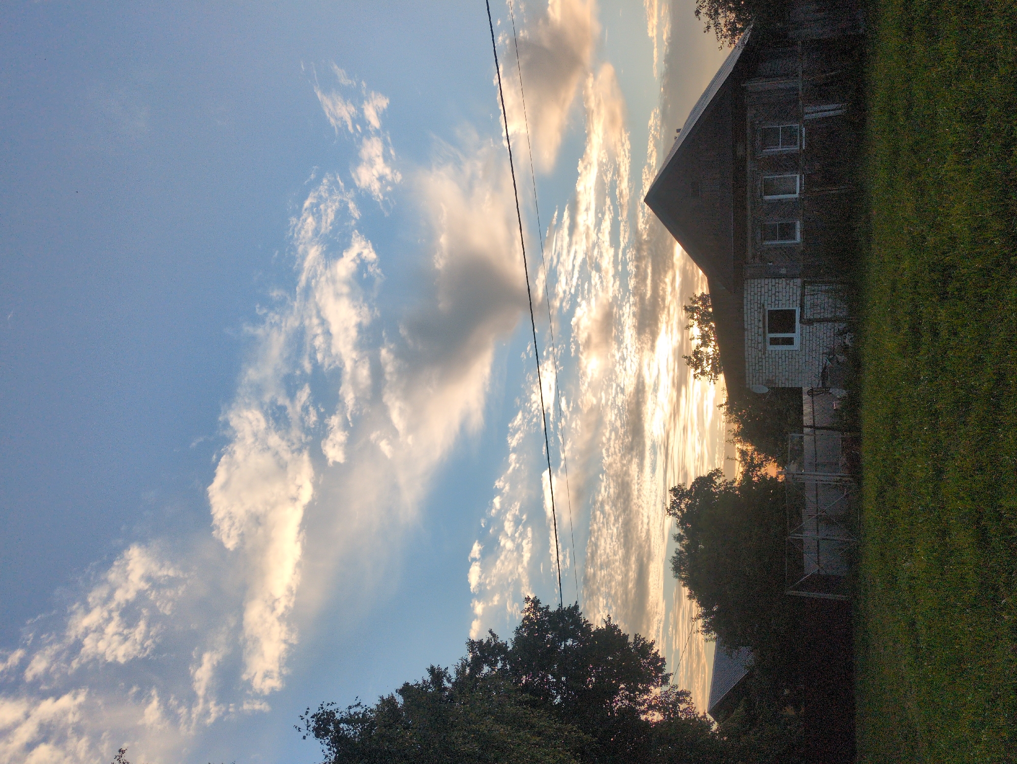 The image depicts a rural scene in Velikovo, Russia, at sunset. The sky is a prominent feature, filled with a mixture of light blue and dramatic clouds illuminated by the setting sun. Streaks of golden and orange light break through the clouds, creating a warm and serene atmosphere. A power line stretches diagonally across the sky, adding a man-made element to the natural landscape.
To the right of the frame, a rustic, two-story house is visible. The lower part of the house appears to be made of white brick, while the upper story is constructed from dark wood. Several windows are visible on the upper floor, and one window is present on the brick portion. The house is partially obscured by dense, dark green trees, which also frame the bottom and right edges of the image, suggesting a natural, somewhat wild setting. A fenced area is also discernible behind the trees, hinting at a yard or garden. The overall impression is one of a peaceful, quiet evening in the countryside. The time of day is clearly sunset, indicated by the warm lighting and the cloud formations. The weather appears clear, with the sunset creating a beautiful spectacle. There is no visible text in the image.