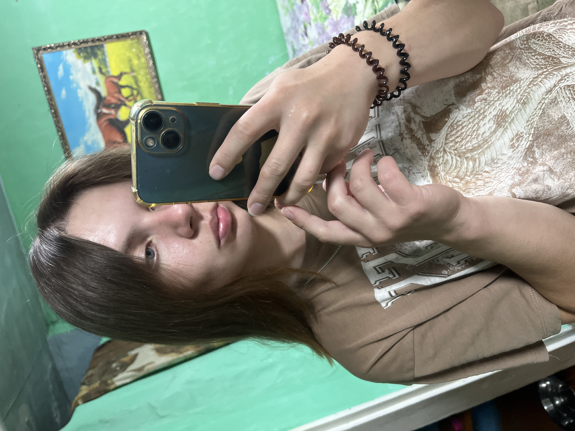 A young person with shoulder-length brown hair is taking a selfie in a room with green walls. They are wearing a tan t-shirt with a graphic design. The person is holding a dark-colored smartphone with a gold case in their right hand. They have two spiral hair ties on their left wrist. In the background, a framed picture of two horses is hanging on the wall. The overall impression is of a casual, indoor setting. There is no indication that this scene is located in Nizhny Tagil, Russia.