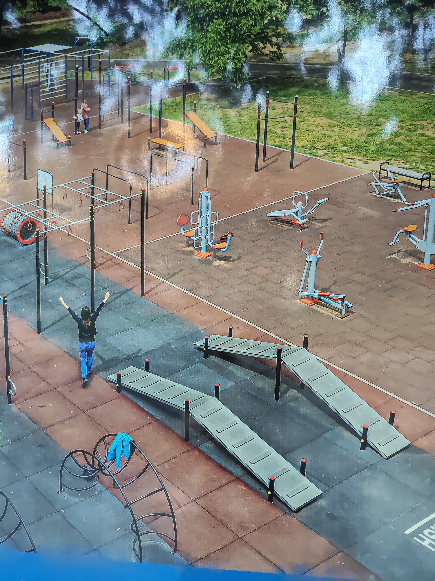 This is an aerial, medium-shot, eye-level photograph of an outdoor public gym in Chișinău, Moldova. The gym is equipped with various exercise machines and structures, including parallel bars, pull-up bars, benches, and balance beams. The ground is covered with rubber tiles in shades of brown and gray. A woman in a dark jacket and blue jeans is in the foreground, facing away from the camera with her arms raised in a celebratory gesture. She is walking on the gray tiled portion of the gym. In the background, there are trees and grass, suggesting the gym is located in a park or a similar green area. The weather appears overcast, with reflections visible on the gym equipment.