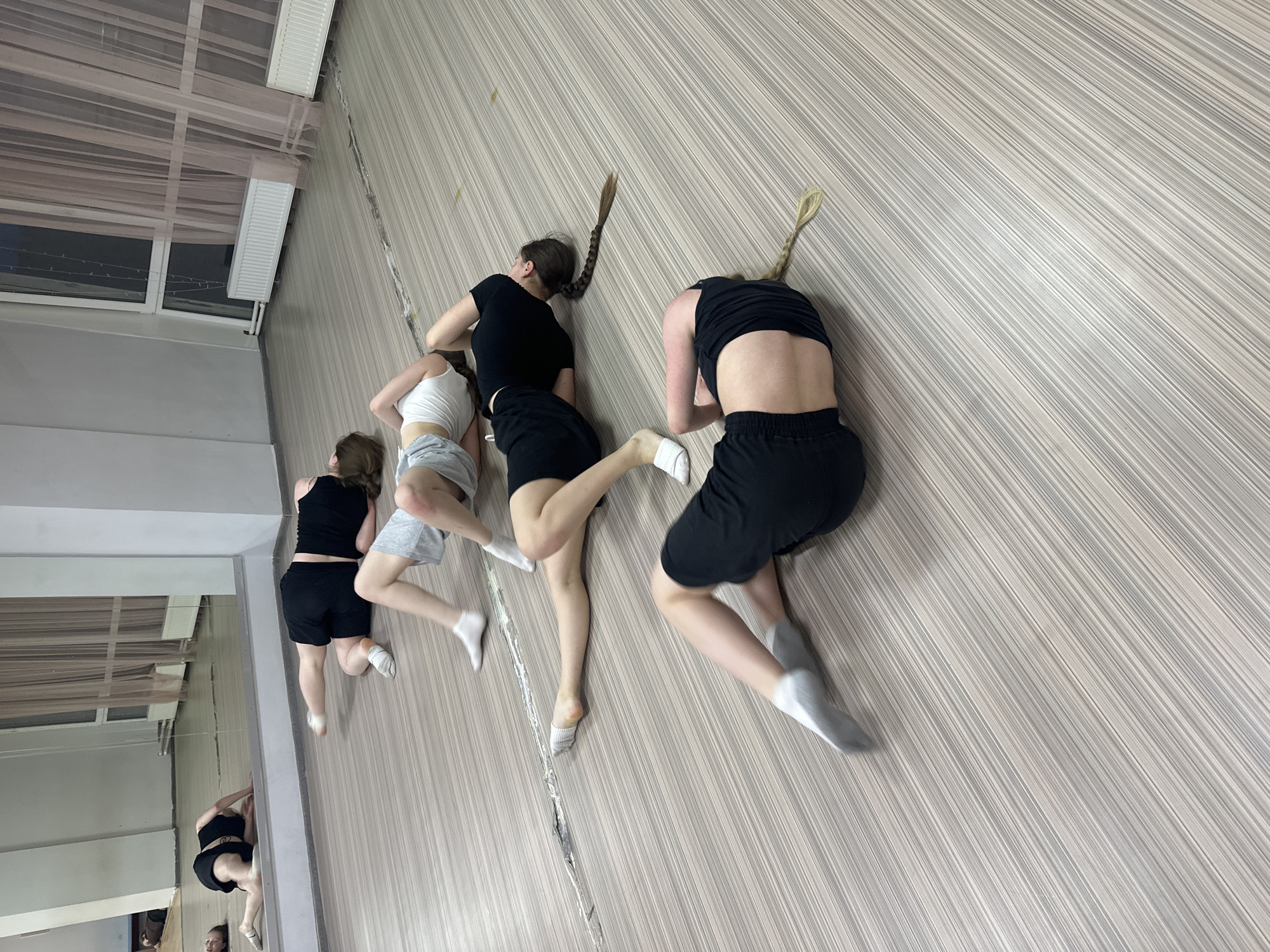 The image depicts an indoor scene, likely a dance or practice studio in Votkinsk, Russia. Four young individuals are resting or posing on a light-colored, striped floor, viewed from an elevated, angled perspective. Most are seen from the back, lying in various prone or supine positions with bent or outstretched limbs, suggesting a choreographed pose or a break during a physical activity like dance or gymnastics.
The individuals are dressed in athletic attire; two wear black shorts and black tops, one in grey shorts with a white crop top, and another in black shorts with a black tank top. Several have long, braided hair. The floor features a distinct pattern of thin, alternating light grey and white stripes, with a prominent seam running diagonally across the surface.
To the left, a large mirror reflects parts of the room, including an additional partially visible person. In the upper background, windows are draped with sheer, light-colored curtains, with white radiators visible beneath them. Fairy lights are strung across the window frames. The room's walls are light-colored, with a vertically striped wallpaper on the right and a plain surface on the left. No text is visible in the image.