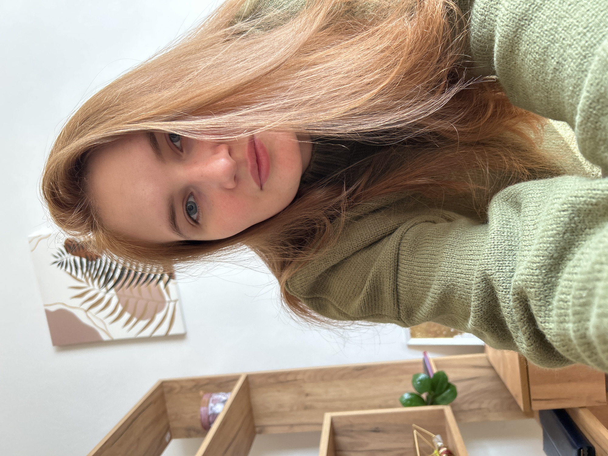 A young woman with fair skin, blue eyes, and light reddish-blonde hair looks at the camera. She is wearing a light green sweater. Behind her is a white wall with a painting of stylized leaves. Below the painting is a wooden shelf with various objects, including a small, green, leafy plant. The location is Chaikovsky, Russia.