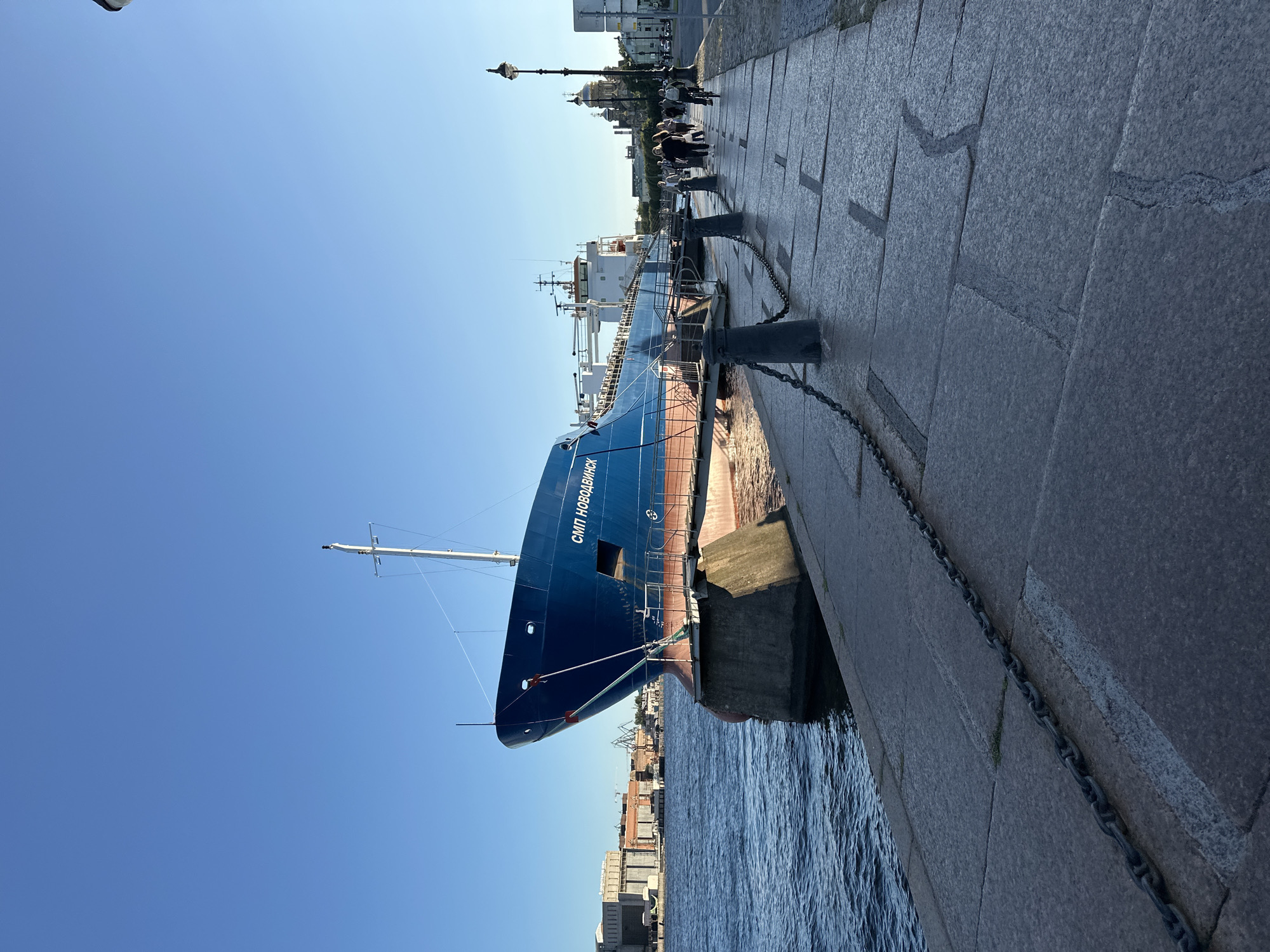 This is a photograph of the St. Petersburg skyline at sunset, taken from the embankment along the Neva River. A large blue cargo ship, identified by the Cyrillic lettering "СМП НОВОДВИНСК" on its side, is docked on the left side of the frame. The ship is anchored to a concrete pier. The embankment is paved with large granite slabs and is lined with black bollards and chains.
Several people are walking along the embankment on the right side of the frame. A woman is riding a bicycle in the background. In the distance, a golden-domed church is visible, along with other buildings that appear to be part of the city's historic architecture. The sky is a clear, vibrant blue, indicating a cloudless day. The lighting suggests it is late afternoon or early evening.
