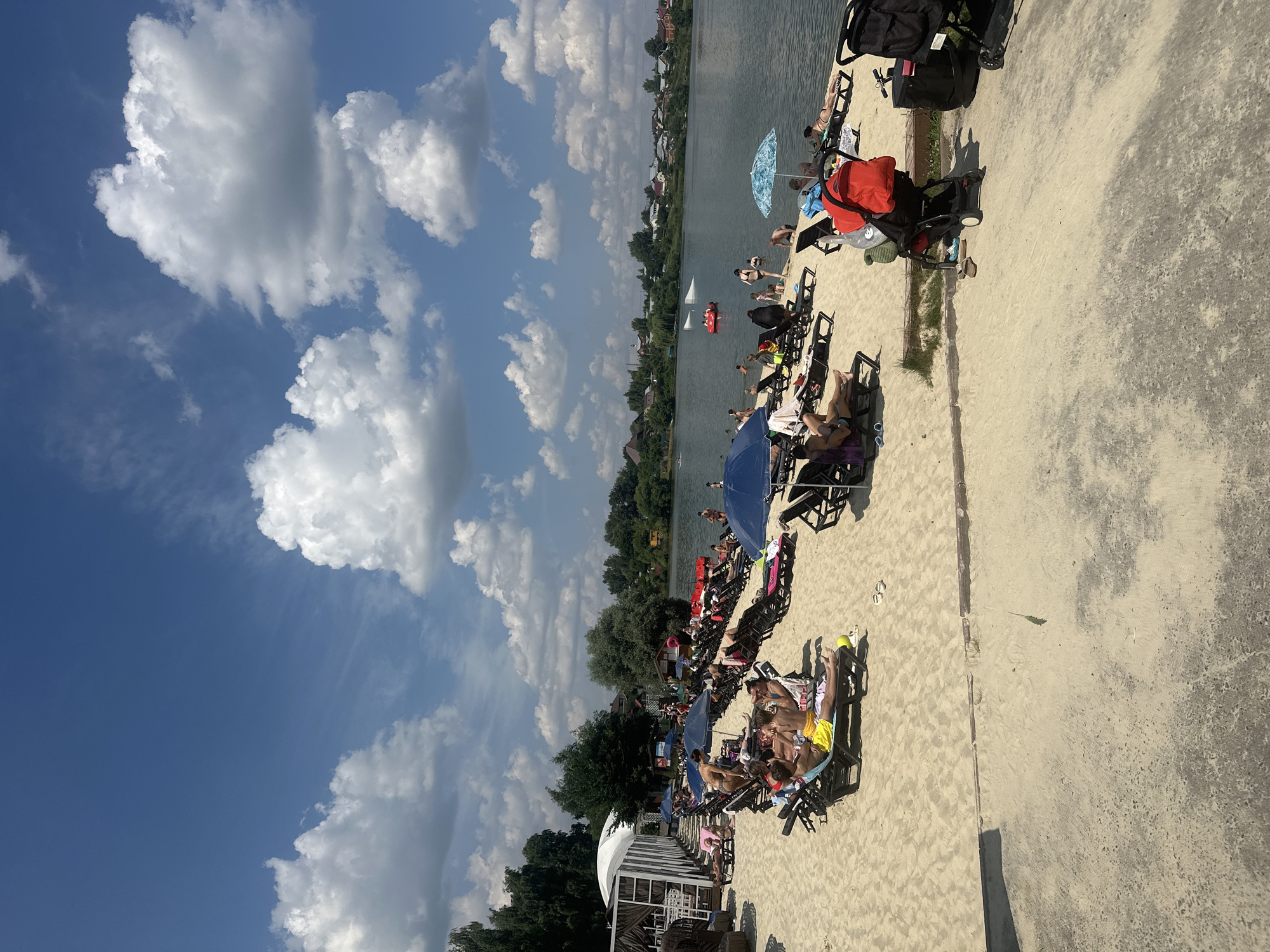 A crowded beach scene unfolds under a partly cloudy sky, with a body of water in the background. The foreground is dominated by sand, with numerous lounge chairs occupied by sunbathers. Umbrellas provide shade over some of the chairs. In the midground, people are seen in the water, some swimming and others on inflatable devices. The background features a shoreline with trees and buildings, suggesting a populated area. The overall impression is a leisurely summer day at a recreational waterfront. This image captures a scene in the city of Molokovo, Russia.