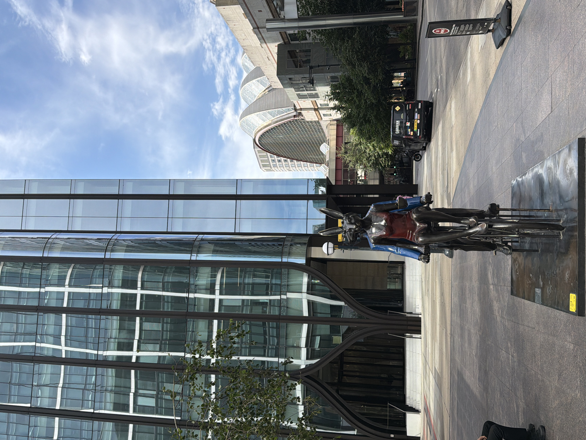 The image depicts a street scene in Canary Wharf, London, United Kingdom. In the foreground, a bronze sculpture of two figures, appearing to be rabbits, riding a tandem bicycle is prominently displayed. The figures are depicted in a dynamic pose, with one wearing a red swimsuit and the other a blue top. The sculpture is situated on a paved area in front of a modern glass building.
The background reveals the distinctive architecture of Canary Wharf, characterized by glass skyscrapers and contemporary structures. To the right, a black van with the word "CARAVAN" visible on its rear is parked on the street, and a red and white sign is placed on the pavement. Trees line the street, and the sky is a clear blue with scattered white clouds. The overall scene suggests an urban environment with a focus on modern architecture and public art.