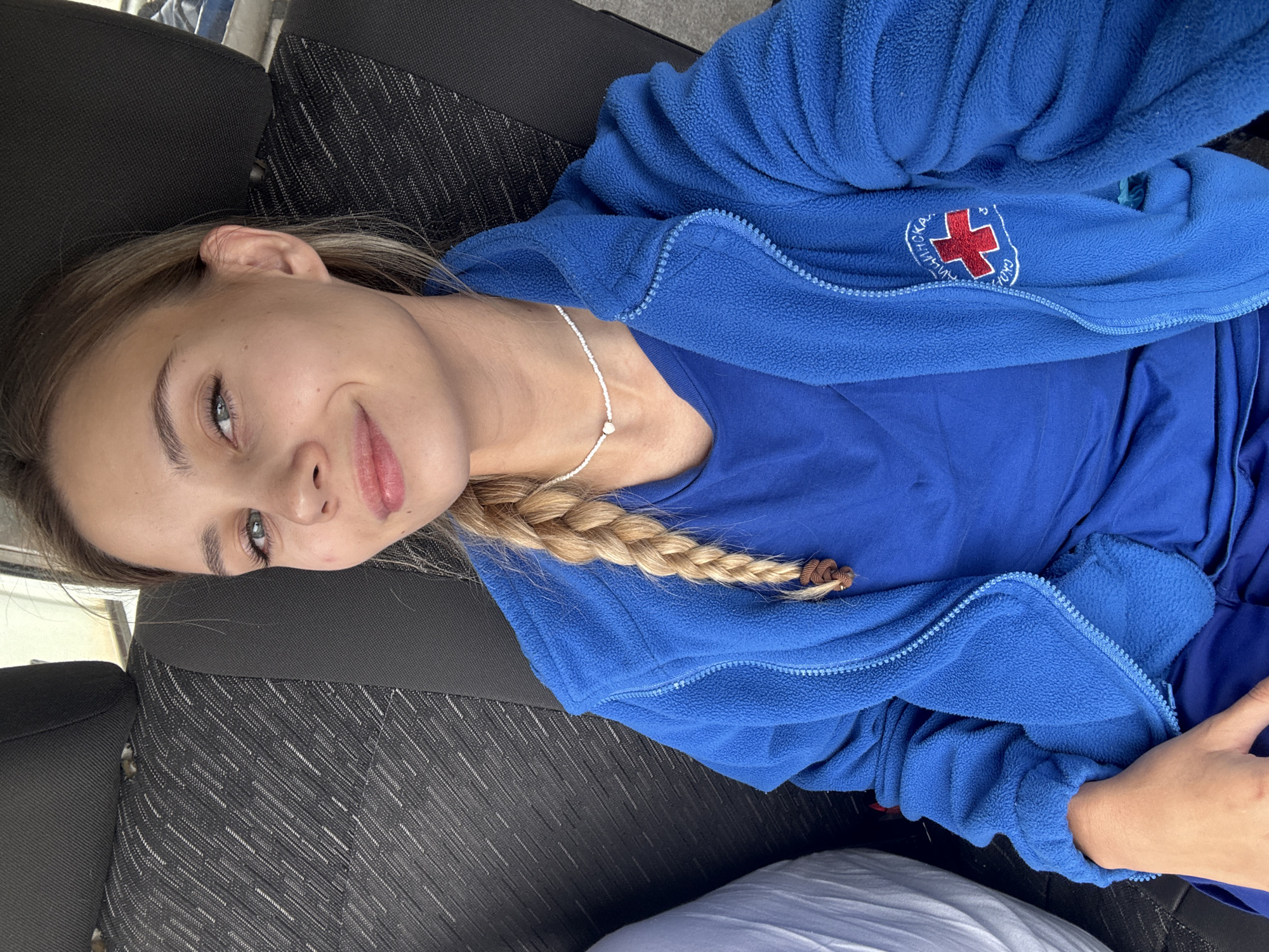 A young adult female is depicted in a close-up, head-and-shoulders shot, seated in the passenger side of a vehicle within the city of Сосново, Russia. She has light blonde hair styled in a single braid over her left shoulder, secured with a brown hair tie. Her eyes are light-colored, and she displays a slight, closed-mouth smile while looking directly at the camera. She is wearing a royal blue fleece zip-up jacket over a plain royal blue t-shirt or scrub top, with dark blue trousers visible below. Around her neck, she wears a white beaded necklace with a small heart-shaped bead in the center. On the left sleeve of her fleece jacket, an embroidered emblem features a red cross on a white background, partially surrounded by Cyrillic text. Visible portions of the text read "КЛИНИЧЕСКАЯ" (Klinicheskaya, meaning "Clinical") above the cross, and "СКОРАЯ" (Skoraya, meaning "Emergency" or "Ambulance") below it, indicating a medical or emergency services uniform. Her right hand rests on her lap. The vehicle's interior features dark grey/black fabric upholstery with a repeating diagonal pattern of small white dots or lines on the seat and headrest behind her. A light-colored fabric or object is visible to her left, beyond the seat.