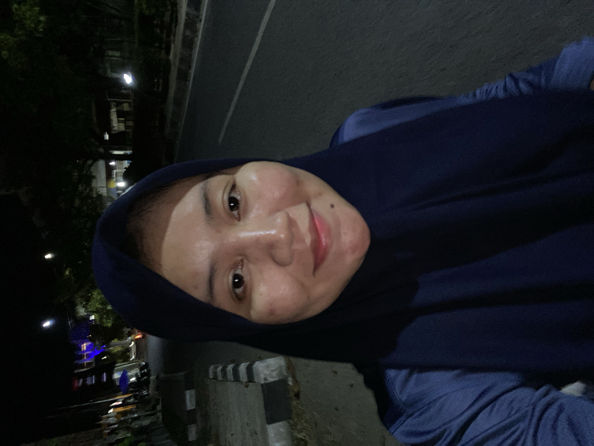 An individual wearing a dark blue hijab and matching long-sleeved shirt is captured in a close-up selfie outdoors at night, on a paved road with a sidewalk and vegetation visible in the background. The individual's face is illuminated, showing clear skin, light brown eyes, and a slight smile. The background suggests an urban or suburban setting, with streetlights and some buildings visible in the distance, characteristic of a street in Banjarbaru, Indonesia. The lighting indicates it is nighttime, likely after dark.