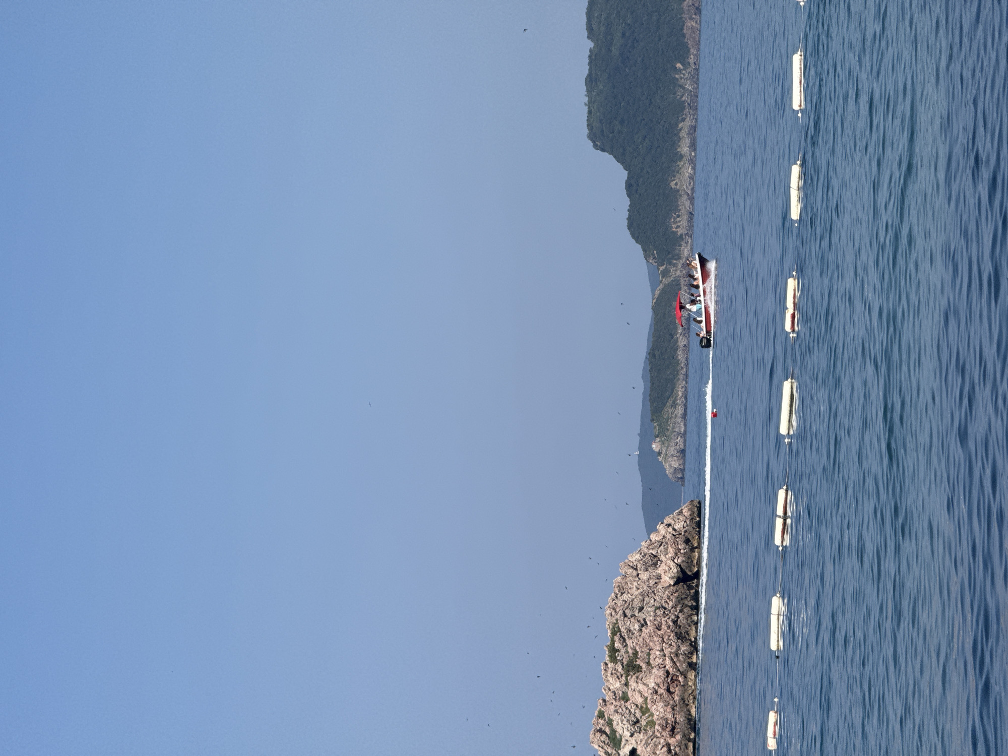 The image captures a bright, sunny day at sea, likely Kamenovo, Montenegro. The water is a deep, rich blue, showing gentle ripples and reflecting the clear sky. A small, motorboat with several people on board is moving across the water, leaving a visible wake. The boat appears to have a red canopy or trim.
Several white buoys, connected by a rope, are floating in a line in the water, suggesting a boundary or marker. In the background, a rugged, rocky island or coastline rises from the sea, covered in lush green vegetation. Another, larger, more distant mountain range can be seen further in the distance, partially shrouded in a light haze. The sky is a clear, pale blue, with a few dark specks that appear to be birds in flight. The overall impression is one of a peaceful, scenic day spent outdoors near the coast. The orientation of the image is rotated 90 degrees counterclockwise.