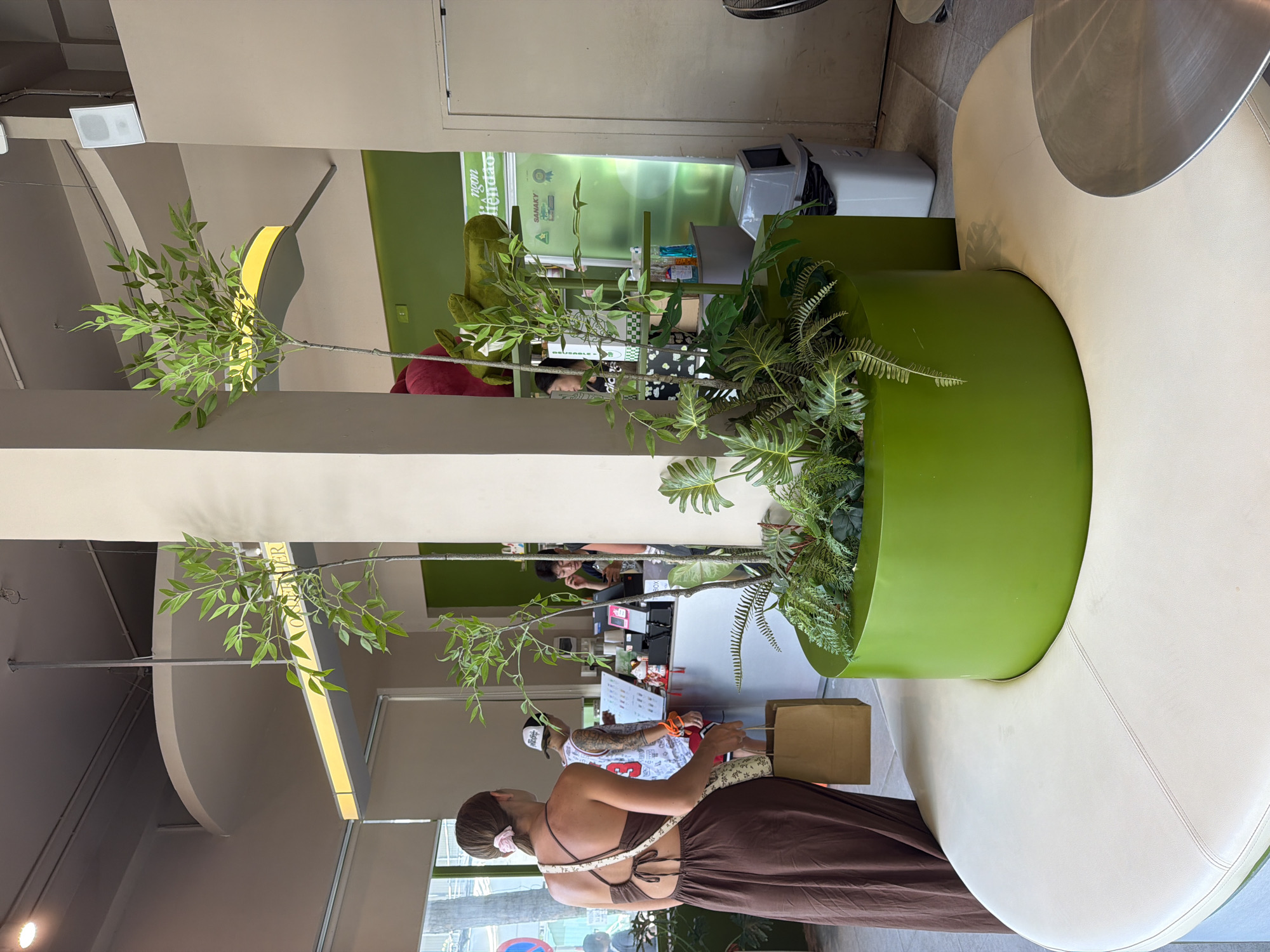 A woman wearing a brown dress stands with her back to the camera in a brightly lit, modern cafe in Nha Trang Ward, Vietnam. She carries a tan paper bag in her left hand. The cafe features a large, circular, cream-colored bench in the foreground, which curves around a tall, vibrant green planter filled with lush foliage. Behind the woman, a counter area is visible where customers are ordering. One man wears a white tank top with red lettering and a black and white baseball cap. To his right, another person, possibly a staff member, is visible behind the counter. The background walls are painted a pale beige and a bright lime green. Modern light fixtures hang from the ceiling, and speakers are mounted on the wall. A silver trash can is situated near the counter. The overall atmosphere is clean and contemporary, with natural elements incorporated through the plants.