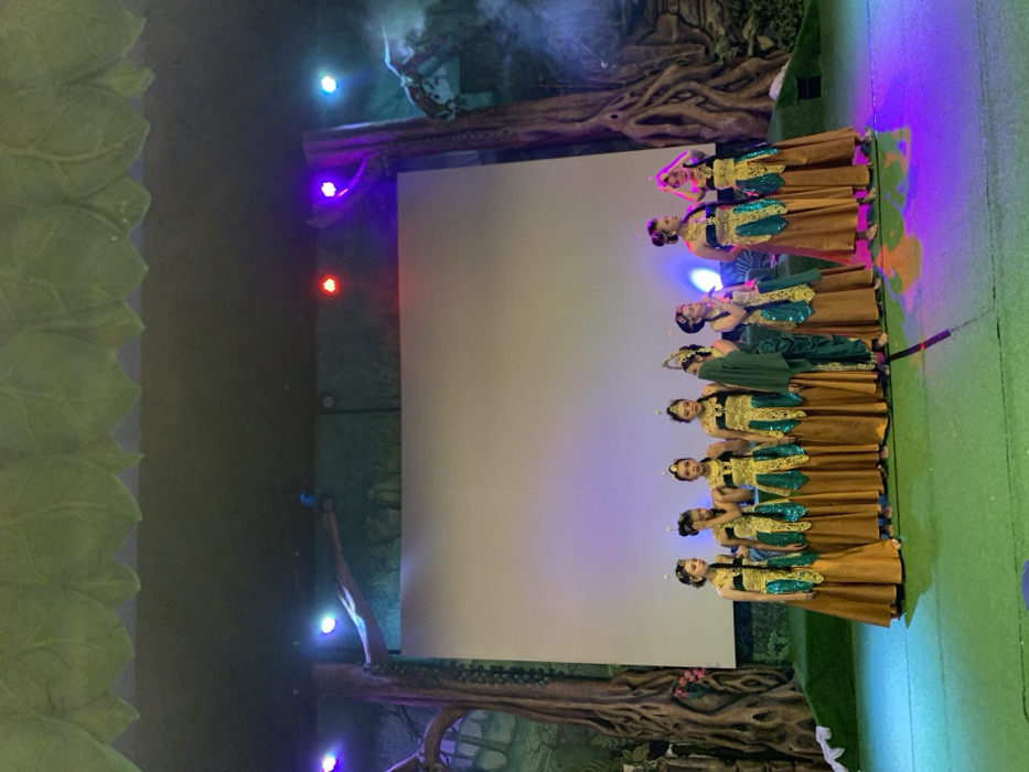 Nine young women are standing on a stage in front of a large white screen. They are wearing gold and green traditional Indonesian costumes and have their hair elaborately styled with flowers. They are all looking at the camera, with some of them smiling. The stage is decorated with fake trees and greenery, and the lighting is dim. The stage is lit with purple and blue lights. This performance is taking place in a theater in Batu, Indonesia at the coordinates -7.885 latitude and 112.524 longitude.
