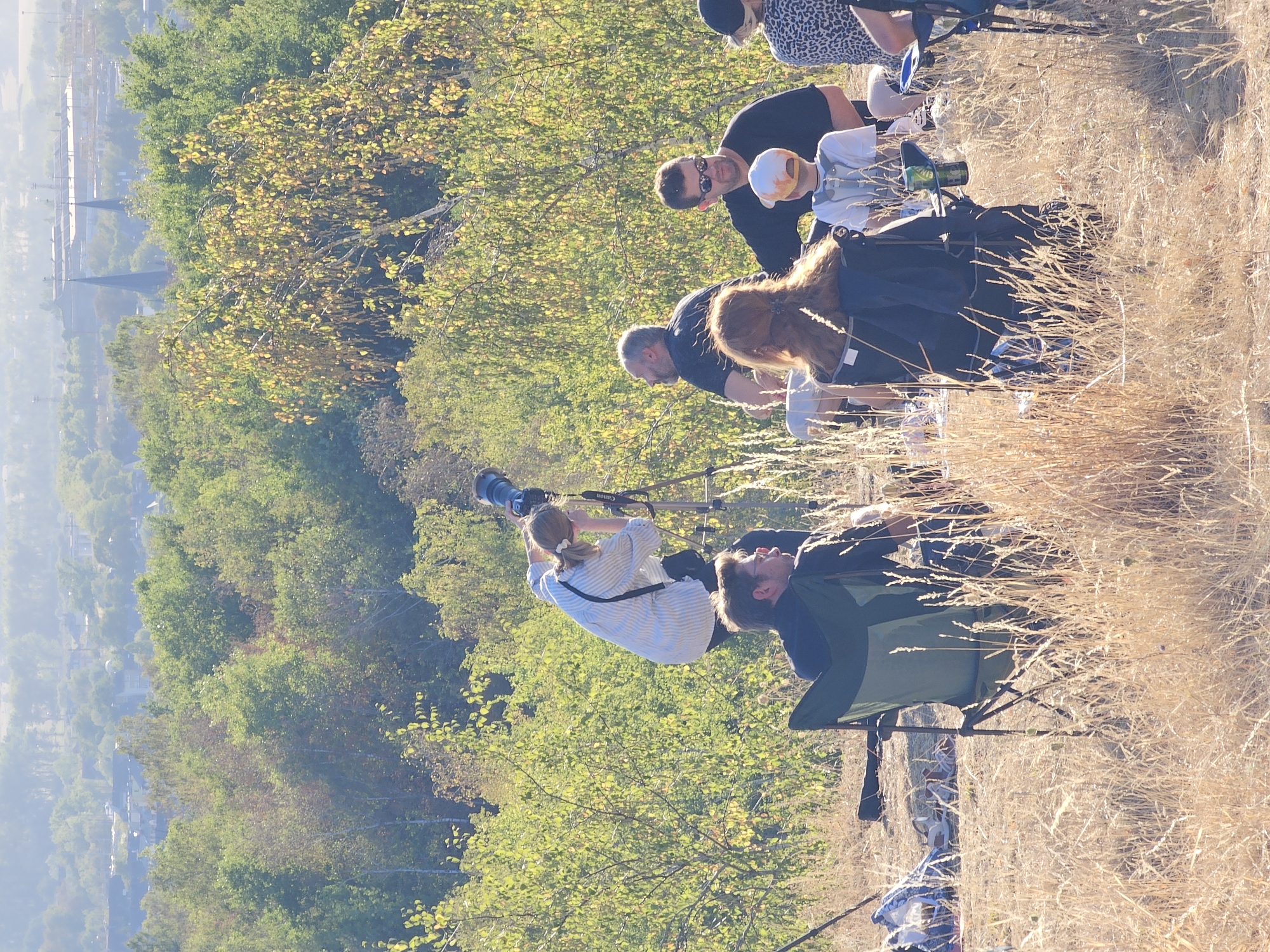 In the city of Schifflange, Luxembourg, a group of individuals is gathered outdoors on a dry, grassy hillside. A woman in a striped shirt is operating a large camera mounted on a tripod, appearing to photograph the landscape. Several other people are seated on camping chairs, facing away from the camera, suggesting they are observing the same vista. One person, a child wearing a hat, is also present in the group. The environment is characterized by dry grass, sparse trees, and a distant view of buildings and industrial structures under a hazy sky, indicating the setting is likely a scenic overlook or a nature area near the town.