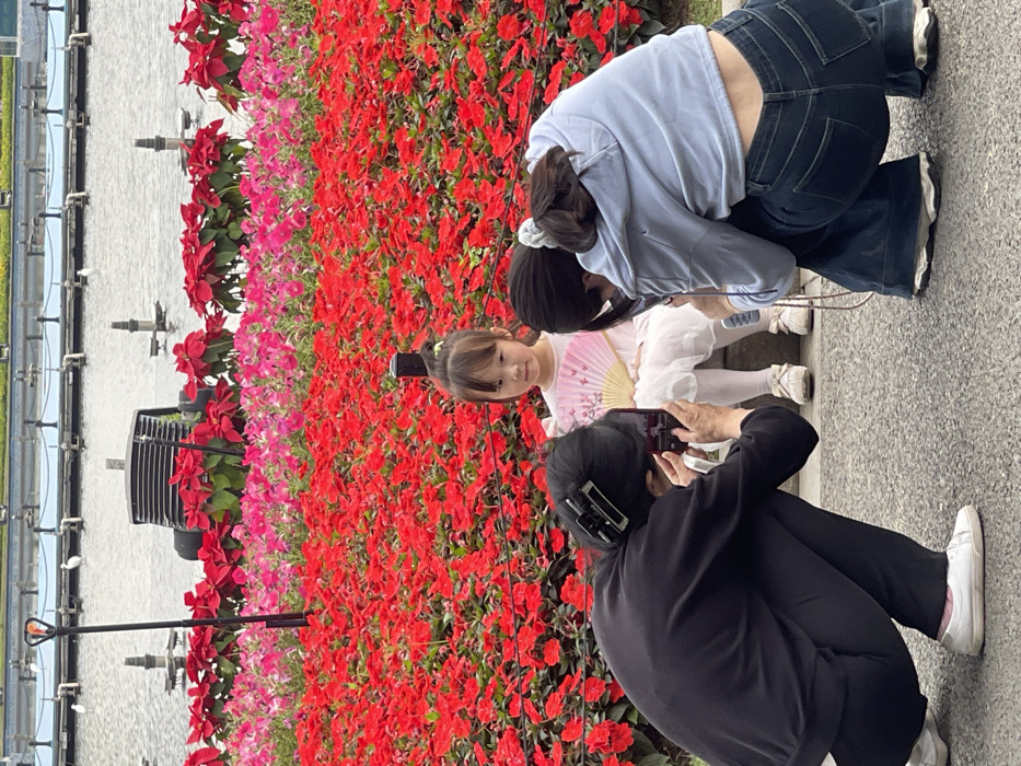 The image depicts a woman in black clothing taking a photograph of a young girl in a white dress and another woman in blue clothing, all standing in front of a wall of red and pink flowers. The location is in Guang Zhou Shi, China, at the coordinates 23.115 latitude and 113.319 longitude. The woman in black clothing is holding a smartphone in her right hand and looking at the screen. The girl is holding a white fan in her left hand and looking at the camera. The woman in blue clothing has her back to the camera, is looking at the girl, and is wearing blue jeans and a light blue sweater. The background is a wall of red and pink flowers.