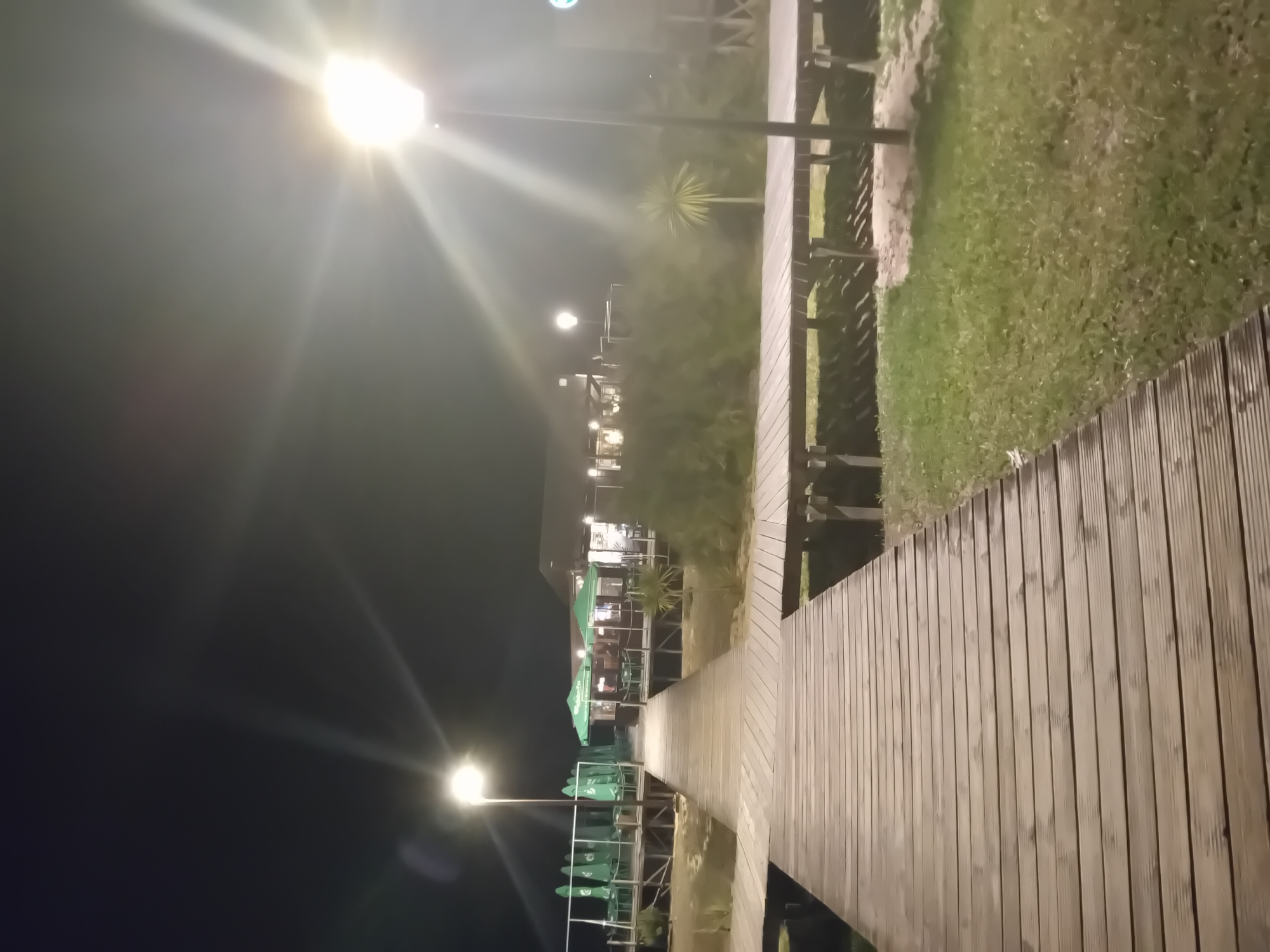 A night-time view of a wooden boardwalk in Figueira da Foz, Portugal. The boardwalk is lit by streetlights, and leads to a building with outdoor seating under green umbrellas. Vegetation, including grass and a small palm tree, borders the boardwalk. The overall atmosphere is dim due to the darkness.